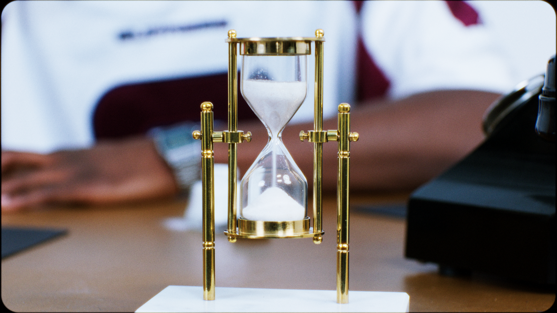 A decorative hourglass with a gold frame and white sand, placed on a desk in an office setting, with blurred people working in the background.