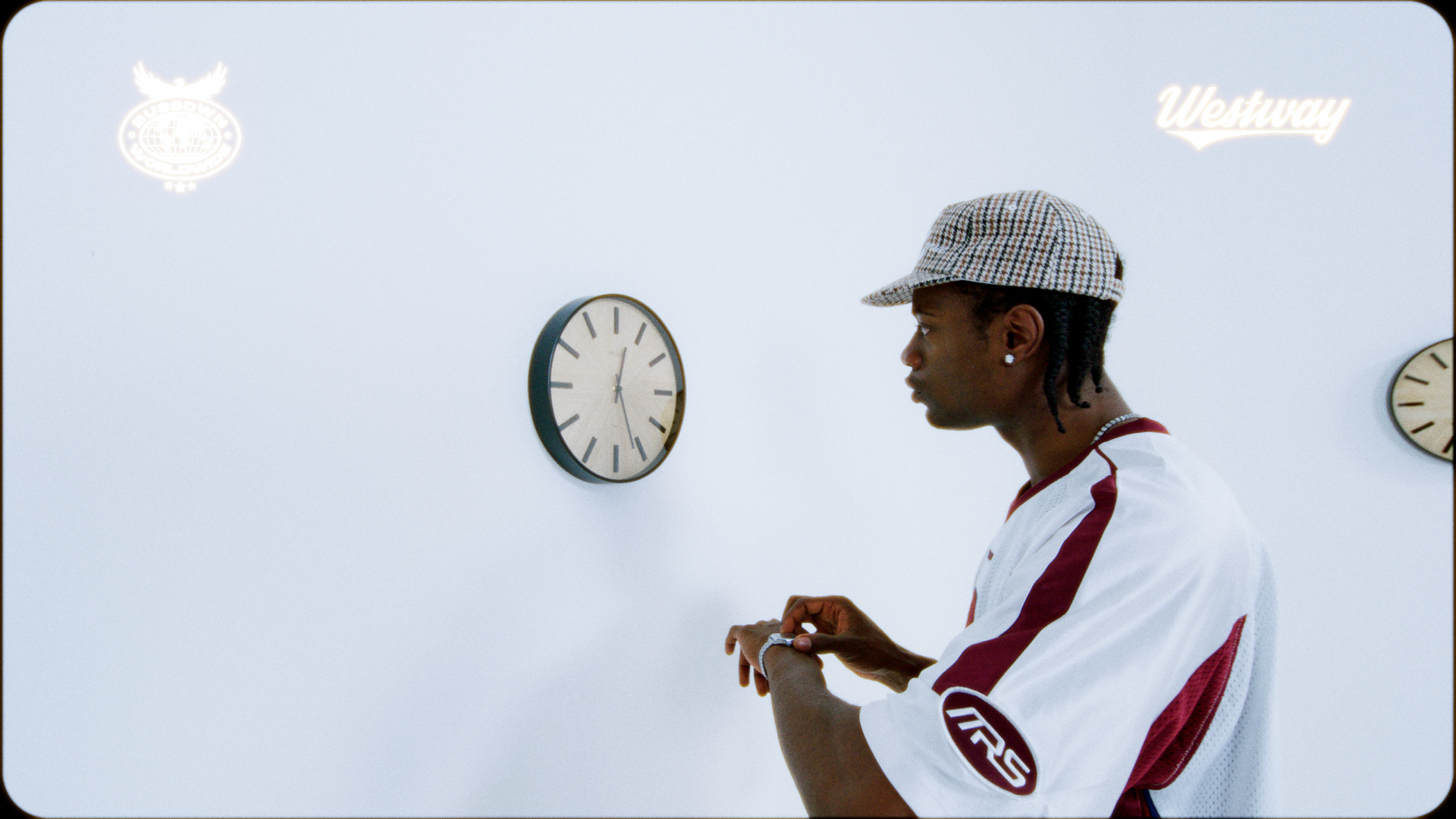 A young man in a white and maroon sports jersey wearing a checkered cap, standing in profile with his hands raised, in front of a white wall with two clocks and illuminated logos reading 'Westway' and a sports brand.