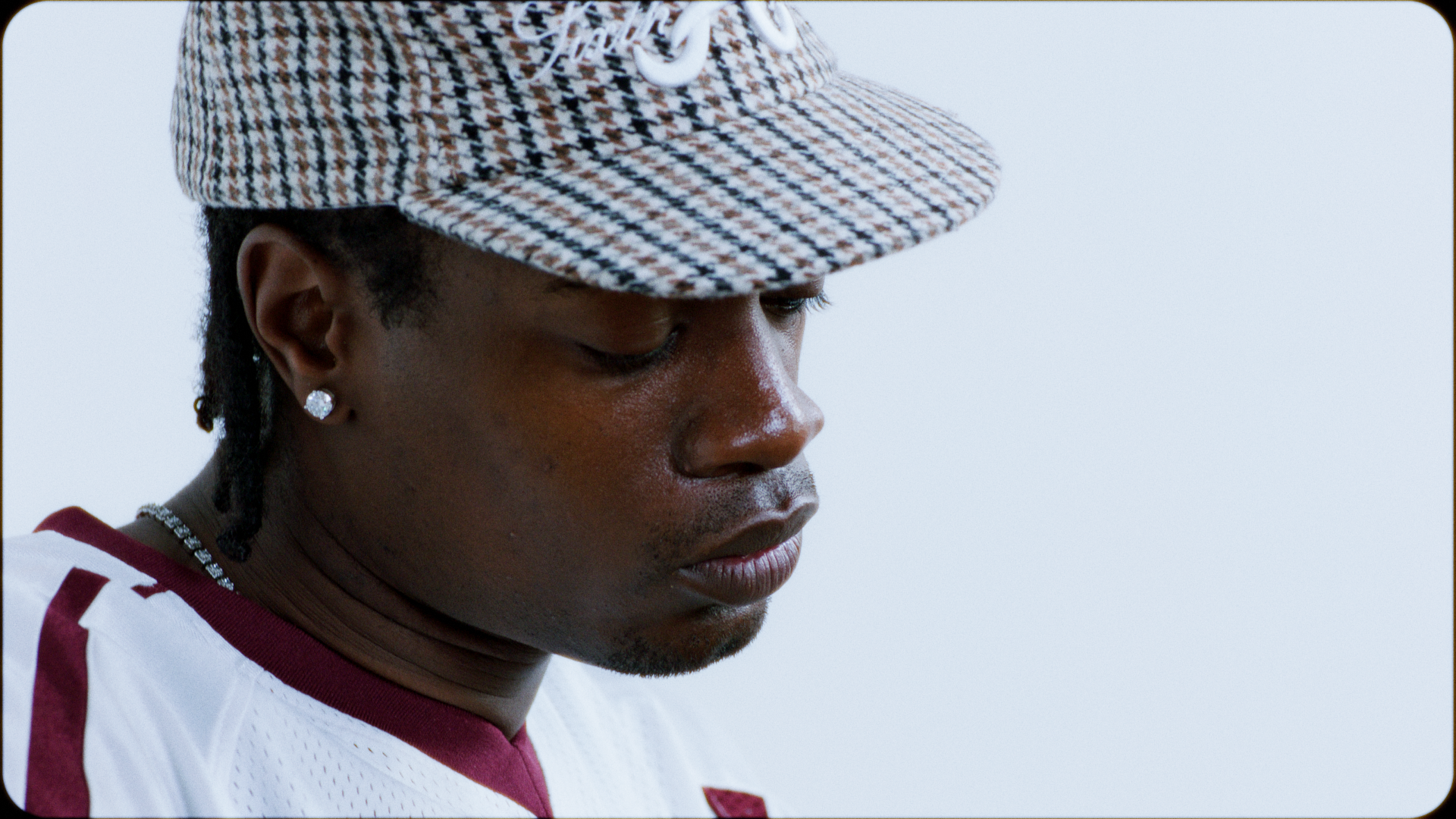 Close-up of a person wearing a checkered baseball cap, earrings, and a white and maroon sports jersey, looking downward against a plain background.