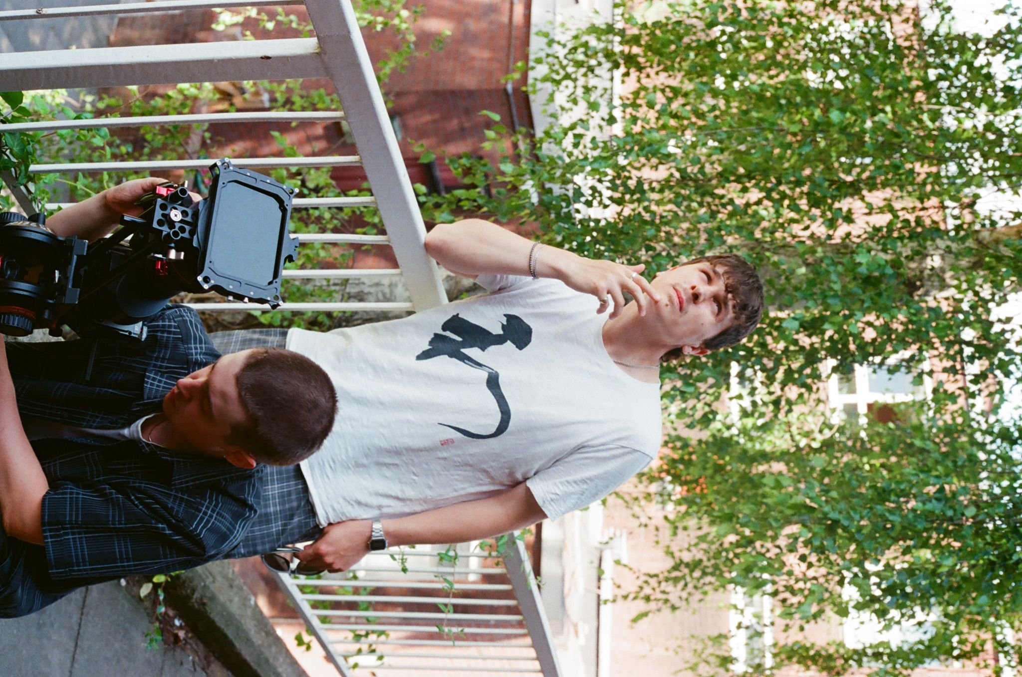 Two young men stand by a metal railing on a balcony or outdoor space with trees and red brick buildings in the background. One man is wearing a white t-shirt with a black graphic, and the other is in a dark checkered shirt. A piece of camera equipment is visible in the foreground.