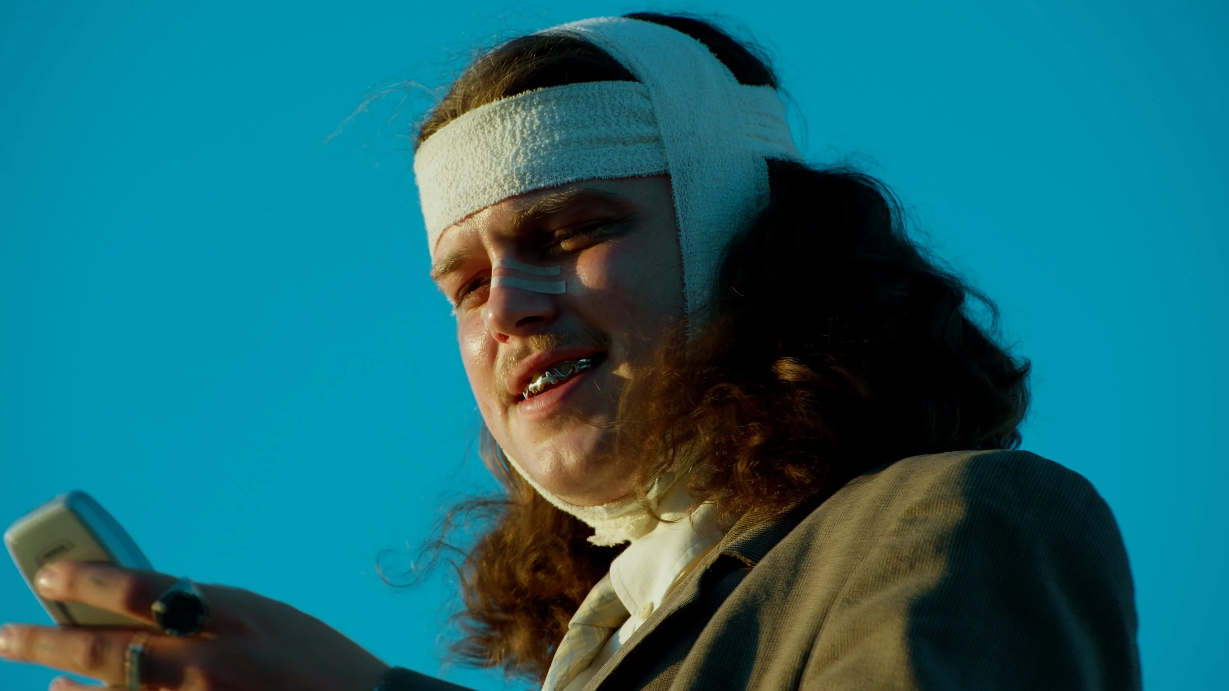 A young person with curly hair, wearing a headband and braces, looks at a smartphone against a blue sky background.