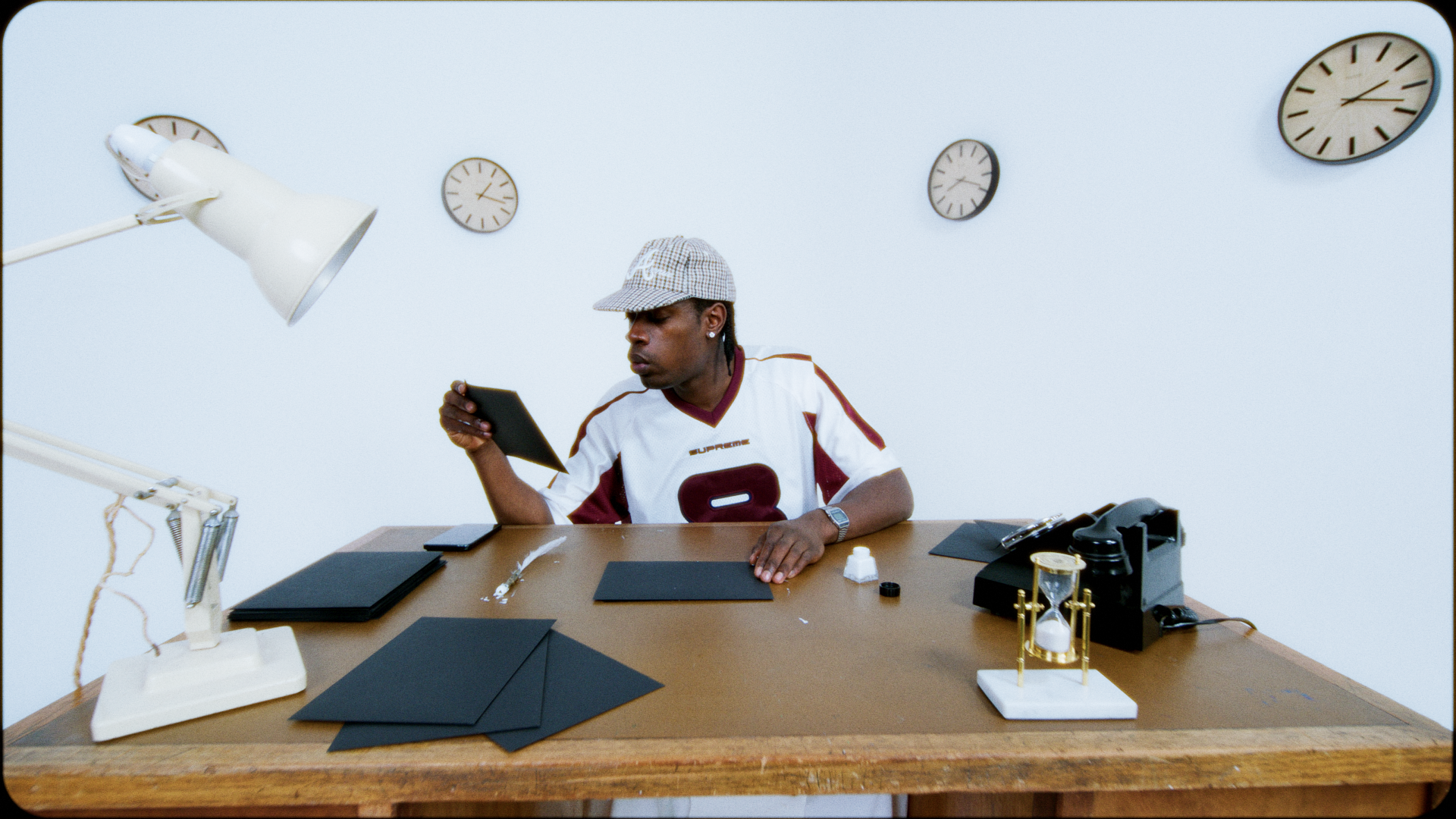 Young man sitting at a desk, looking at his phone in an office or lab setting with clocks on the wall behind him.