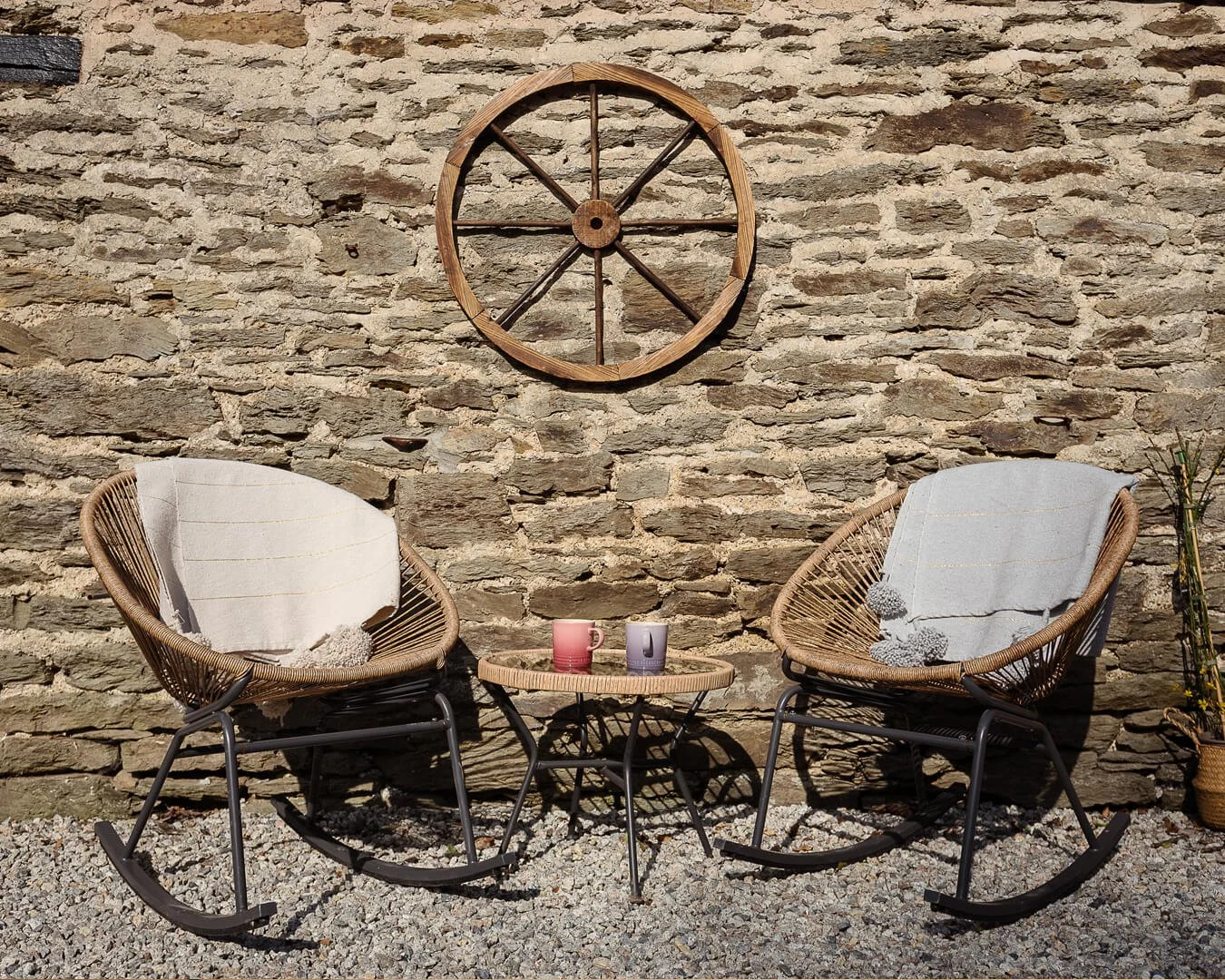 Two wicker rocking chairs with white cushions and soft throws, a small round table with two pastel-colored cups, set against a rustic stone wall at Melancoose, in North Cornwall