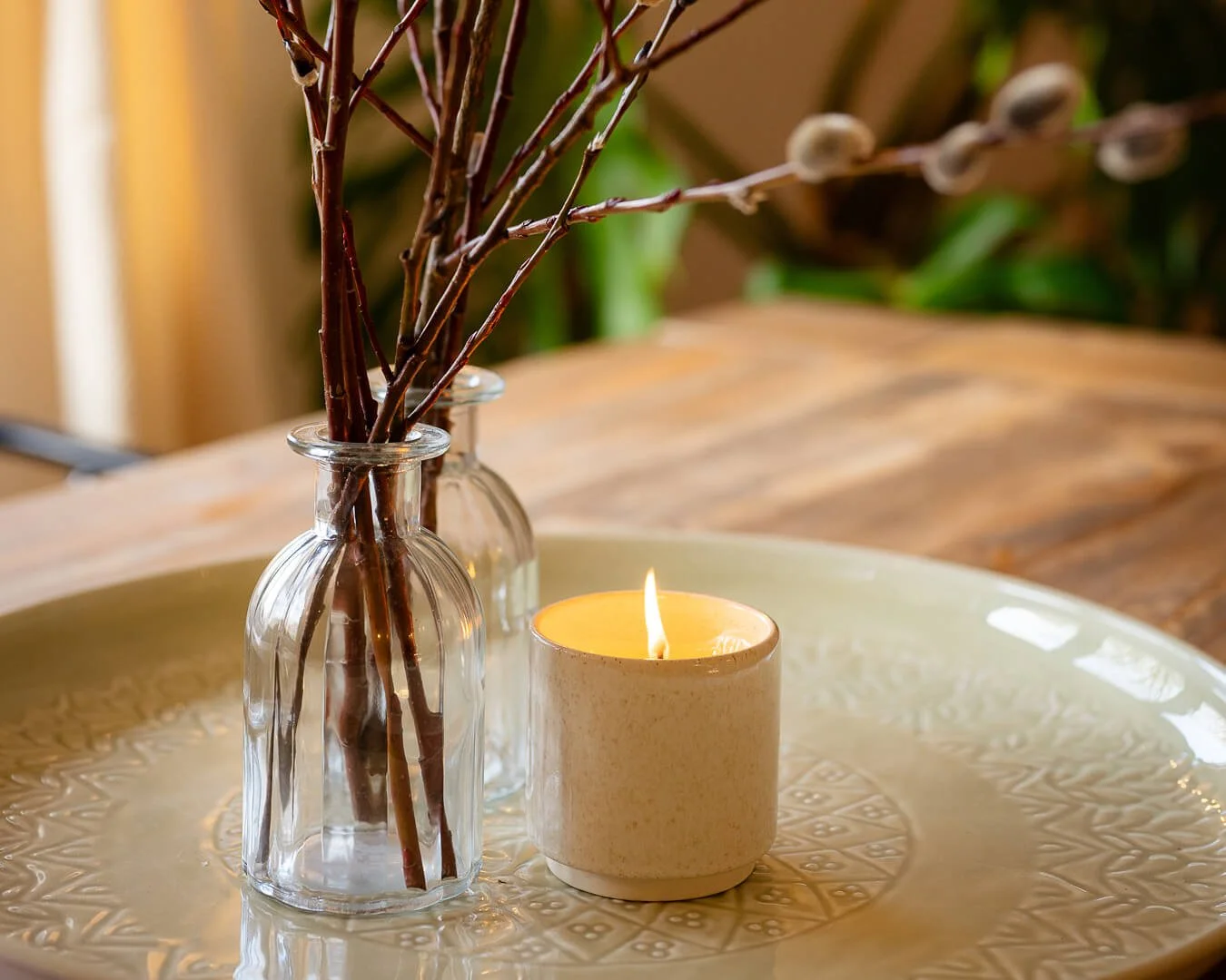 Small candle on a patterned plate with small glass vases of branches in the snug room at Melancoose barn