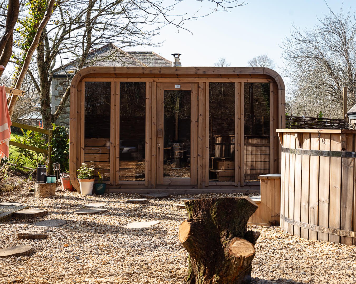A wooden outdoor sauna at Melancoose, with a glass door and windows, surrounded by trees, under a clear sky.