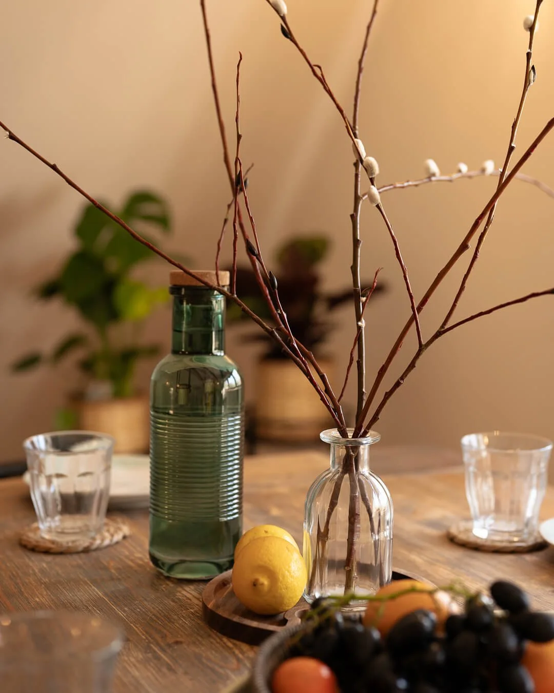 A wooden dining table with a clear glass bottle holding bare branches, a lemon on a wooden coaster, six glasses, a basket of grapes, and potted plants in the background.