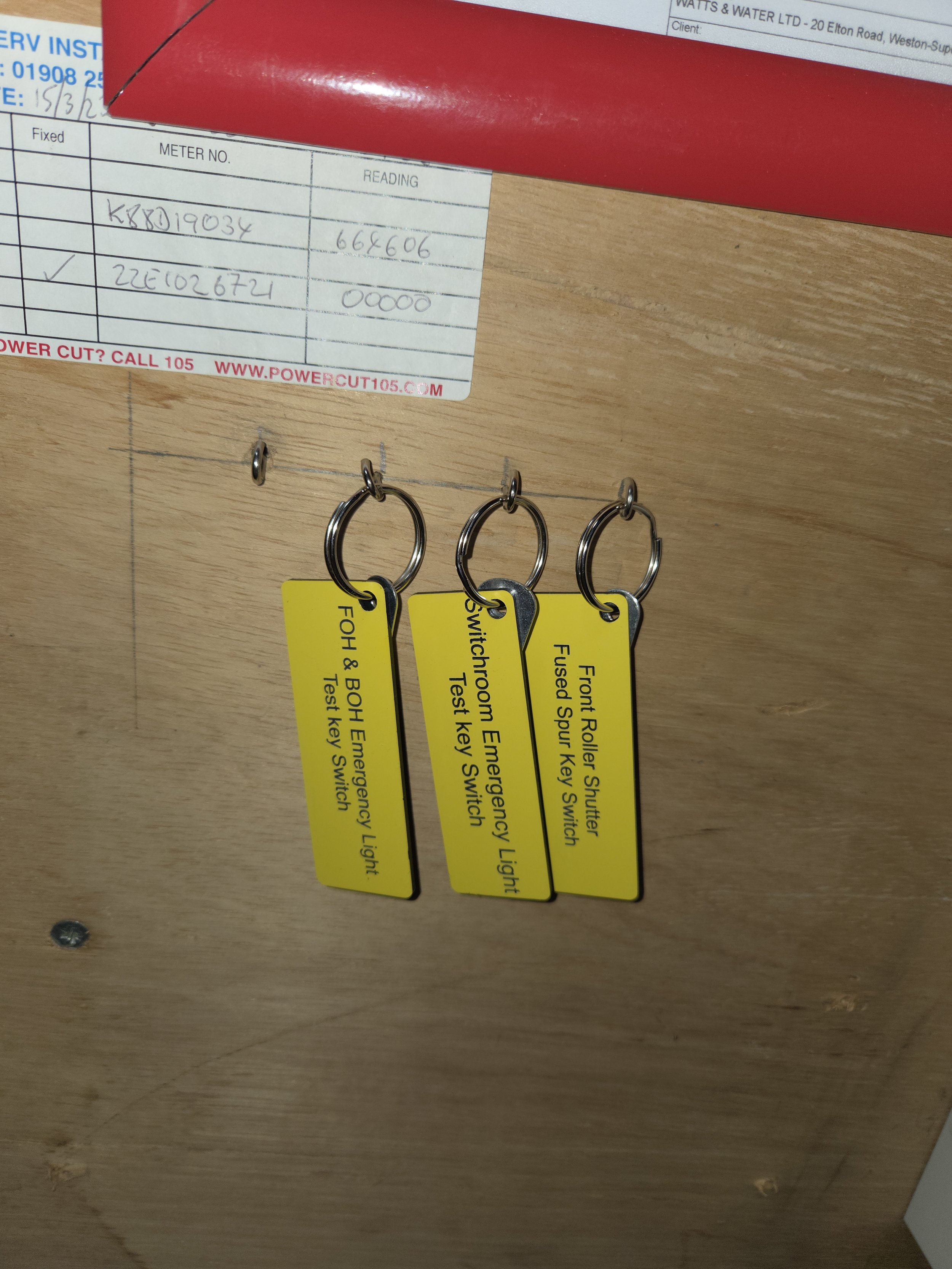 Three yellow tags hanging from hooks, labeled with different switch test instructions: "FOH & BOH Emergency Light Test Key Switch," "Switchroom Emergency Light Test Key Switch," and "Front Roller Shutter Shutter Fused Spur Key Switch."