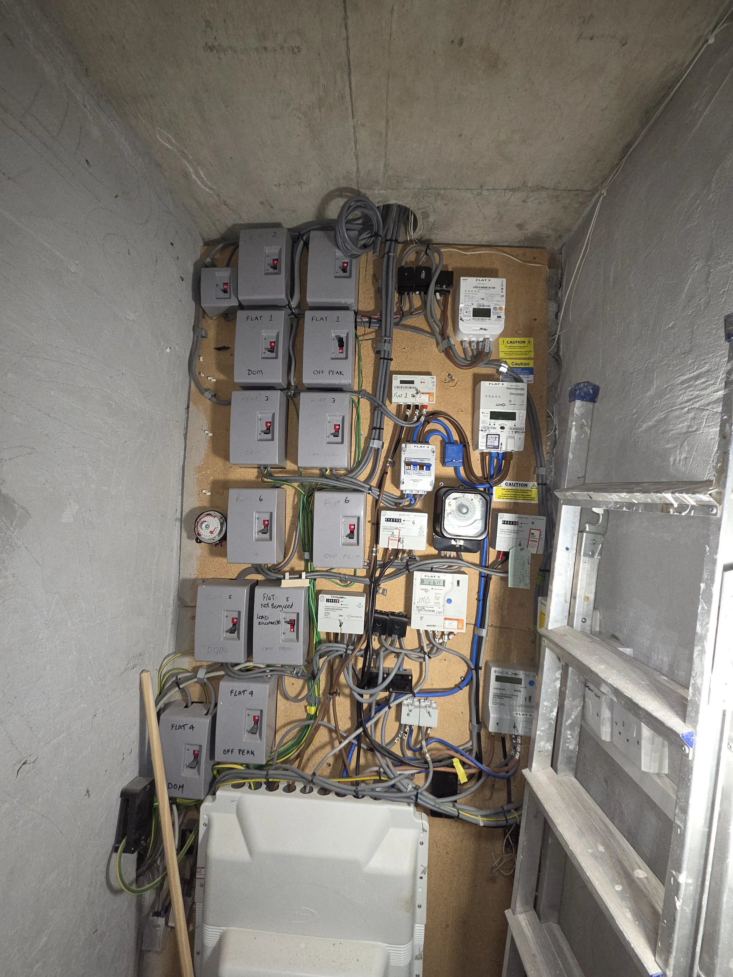 Electrical control panel with various switches, meters, and wiring inside a small unfinished room, with a ladder and a plastic container.