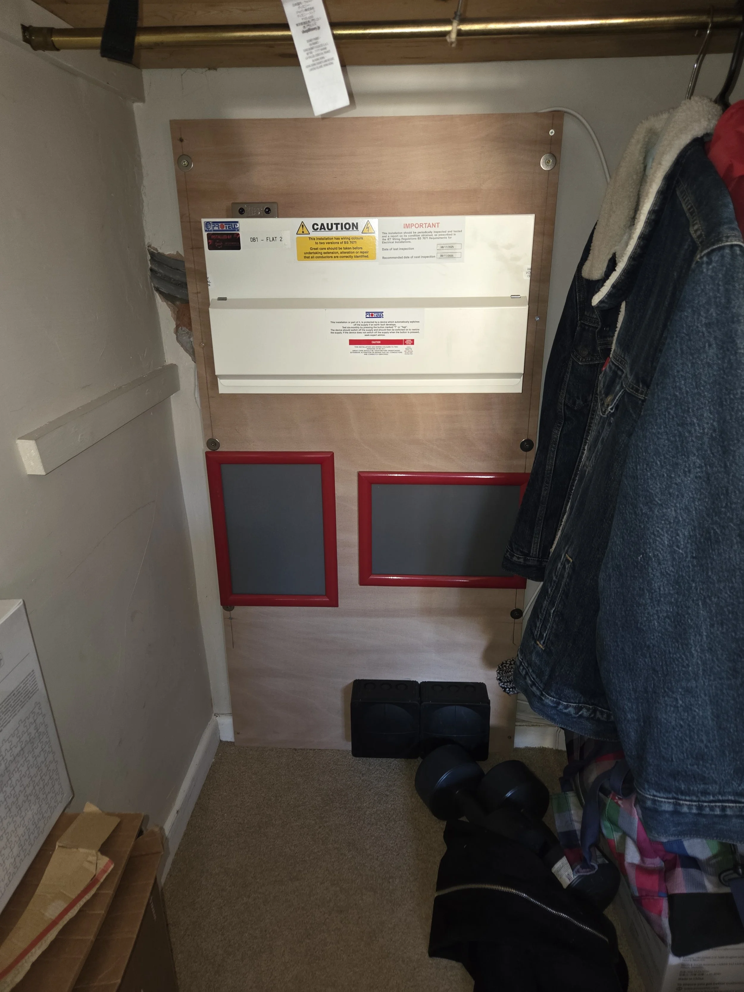 An electrical panel mounted on a wooden board in a small closet with a pair of black speakers and a shoe on the floor, and clothes hanging on a nearby rack.