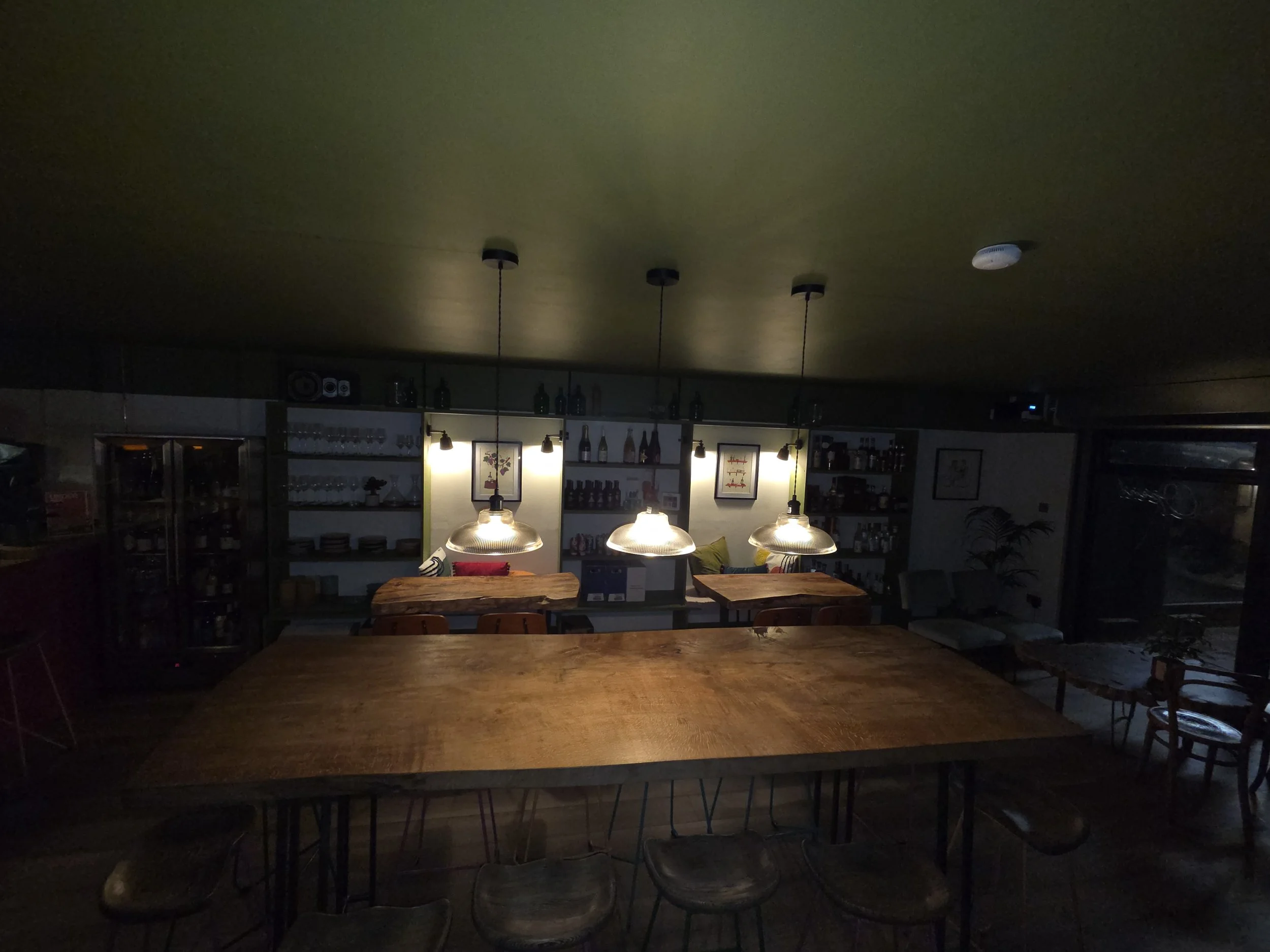 Dimly lit bar or cafe interior with wooden tables, chairs, and pendant lights over the tables, with shelves of bottles and glasses in the background.