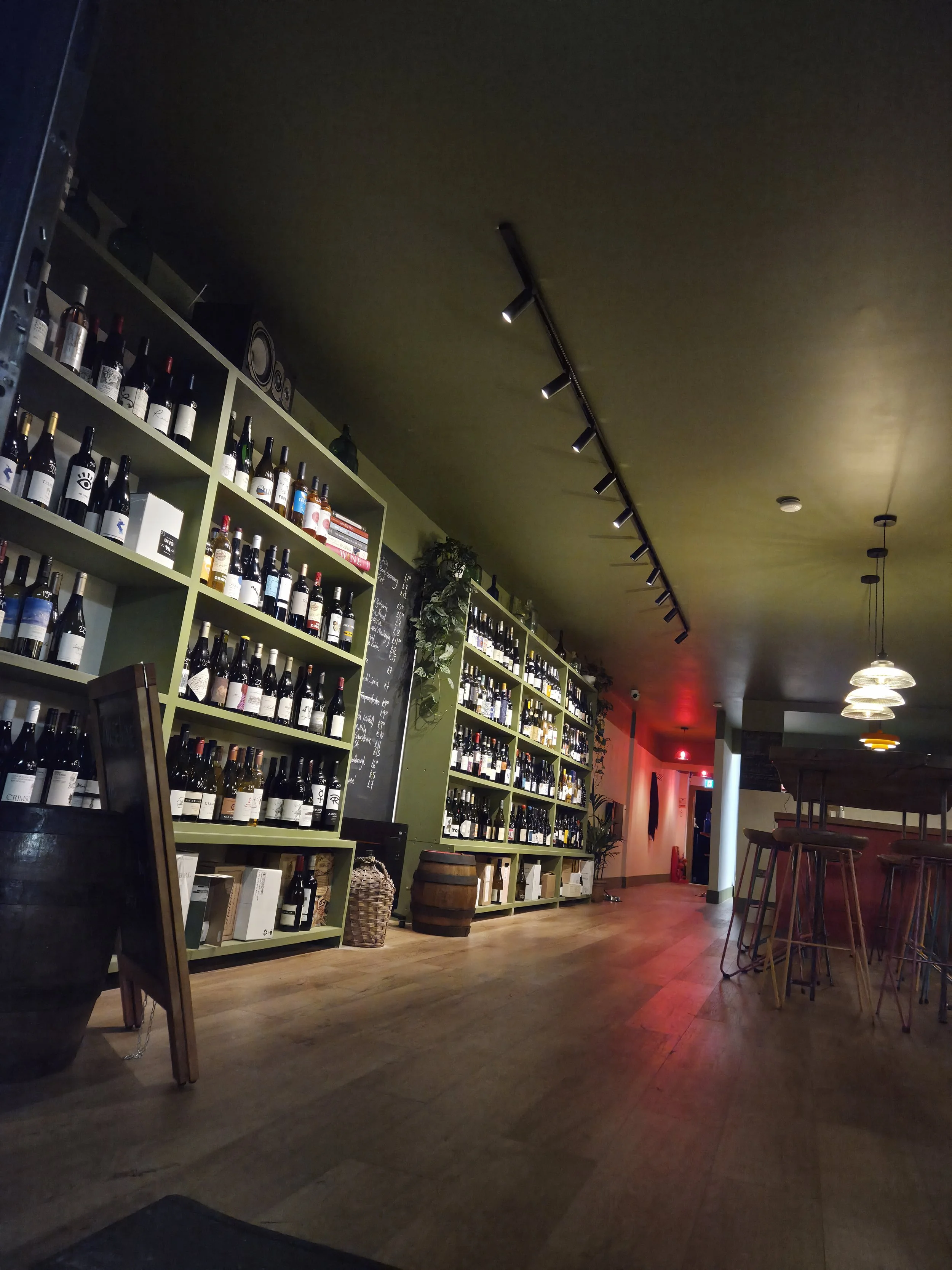 Interior of a wine store or wine bar with green shelves full of wine bottles, a barrel, a chalkboard with writing, and a bar area with high stools and hanging lights.