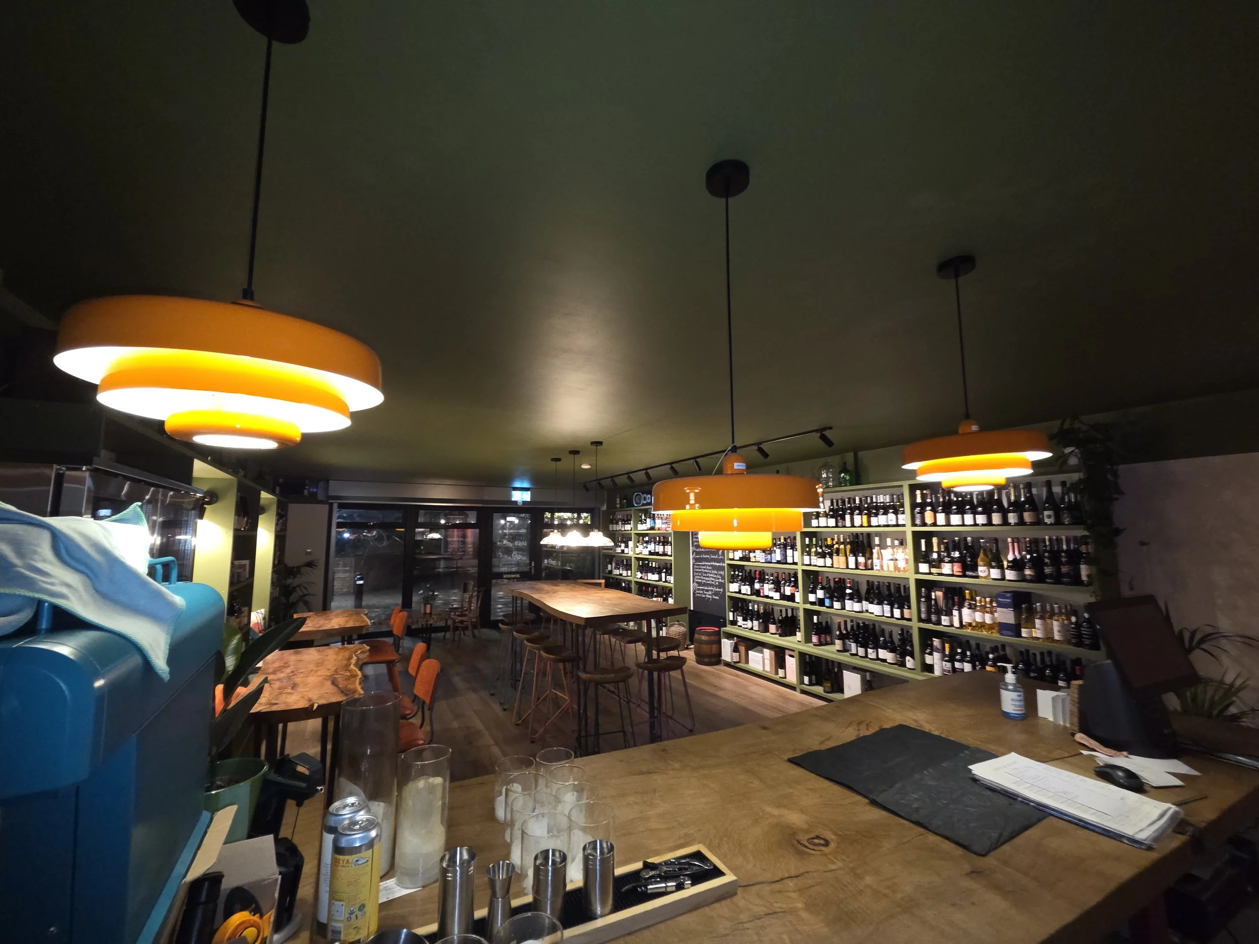 Interior of a cozy bar or restaurant with orange hanging lights, wooden tables and chairs, and shelves stocked with bottles.