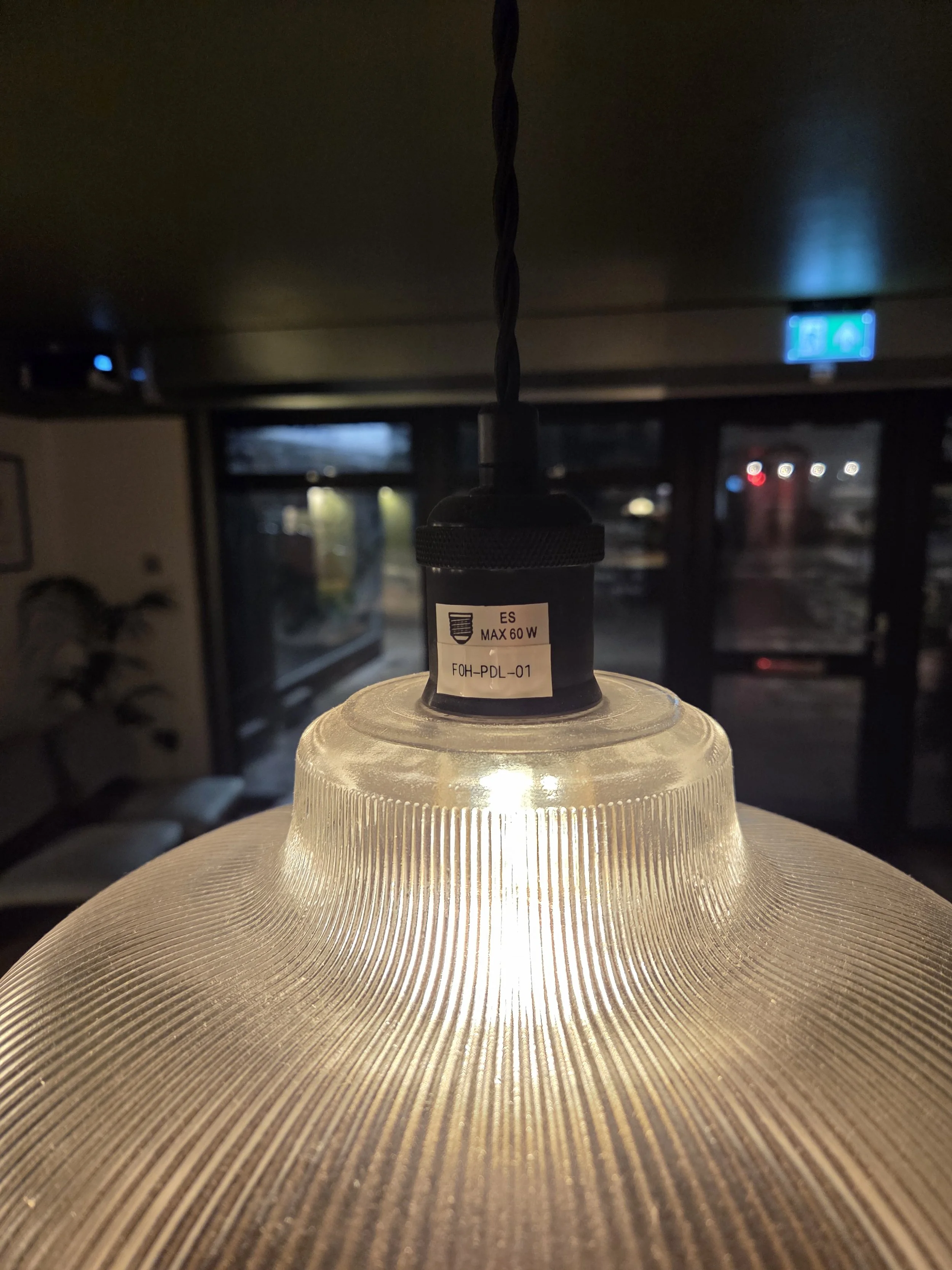 Close-up of a ceiling light fixture with a textured glass cover, hanging from a twisted black cord in a dimly lit interior space. Through the window in the background, some lights and outdoor scenery are visible.
