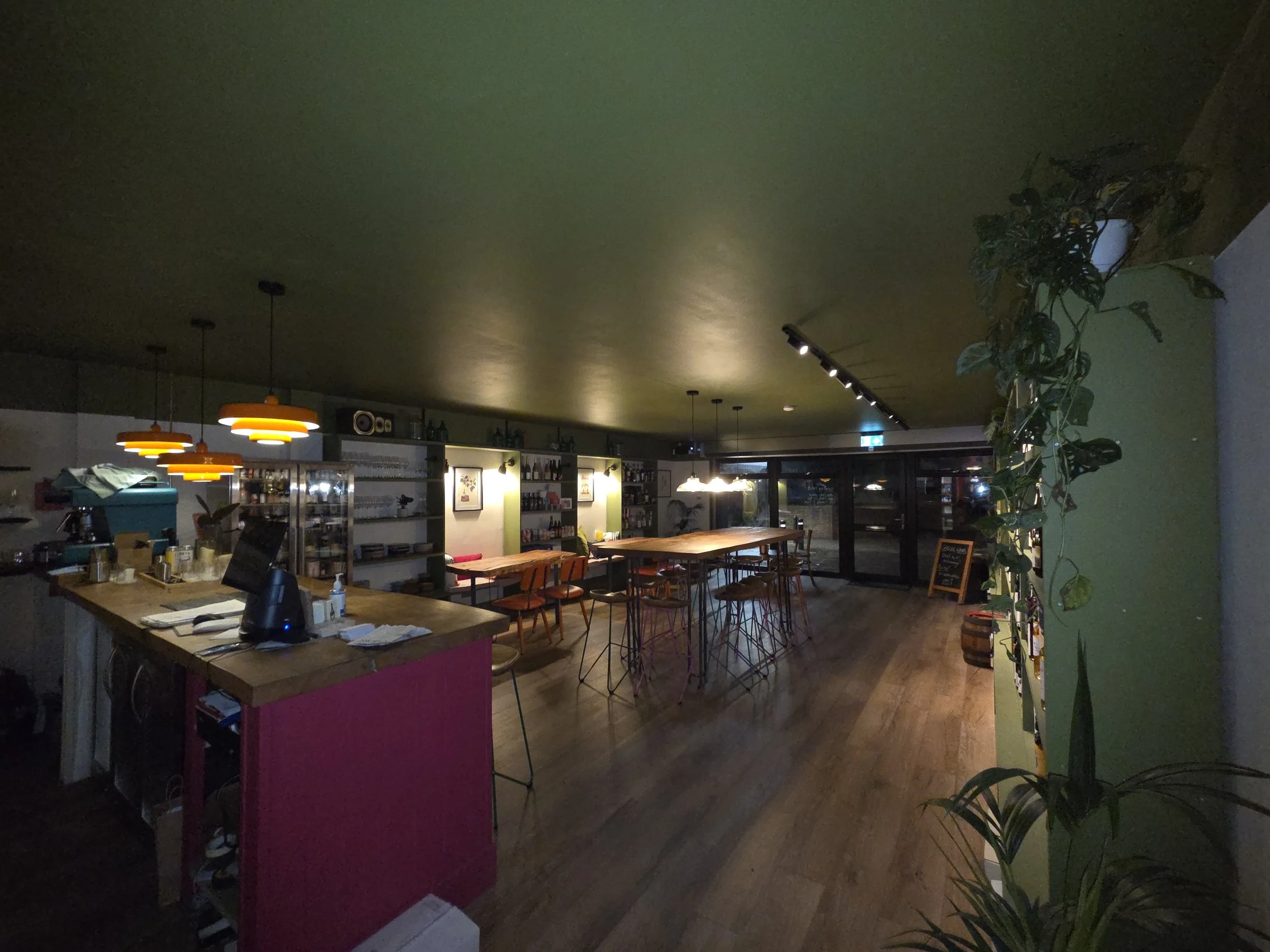 Interior of a cozy cafe with wooden tables and chairs, pendant lighting, and decorative plants along the wall, illuminated softly at night.