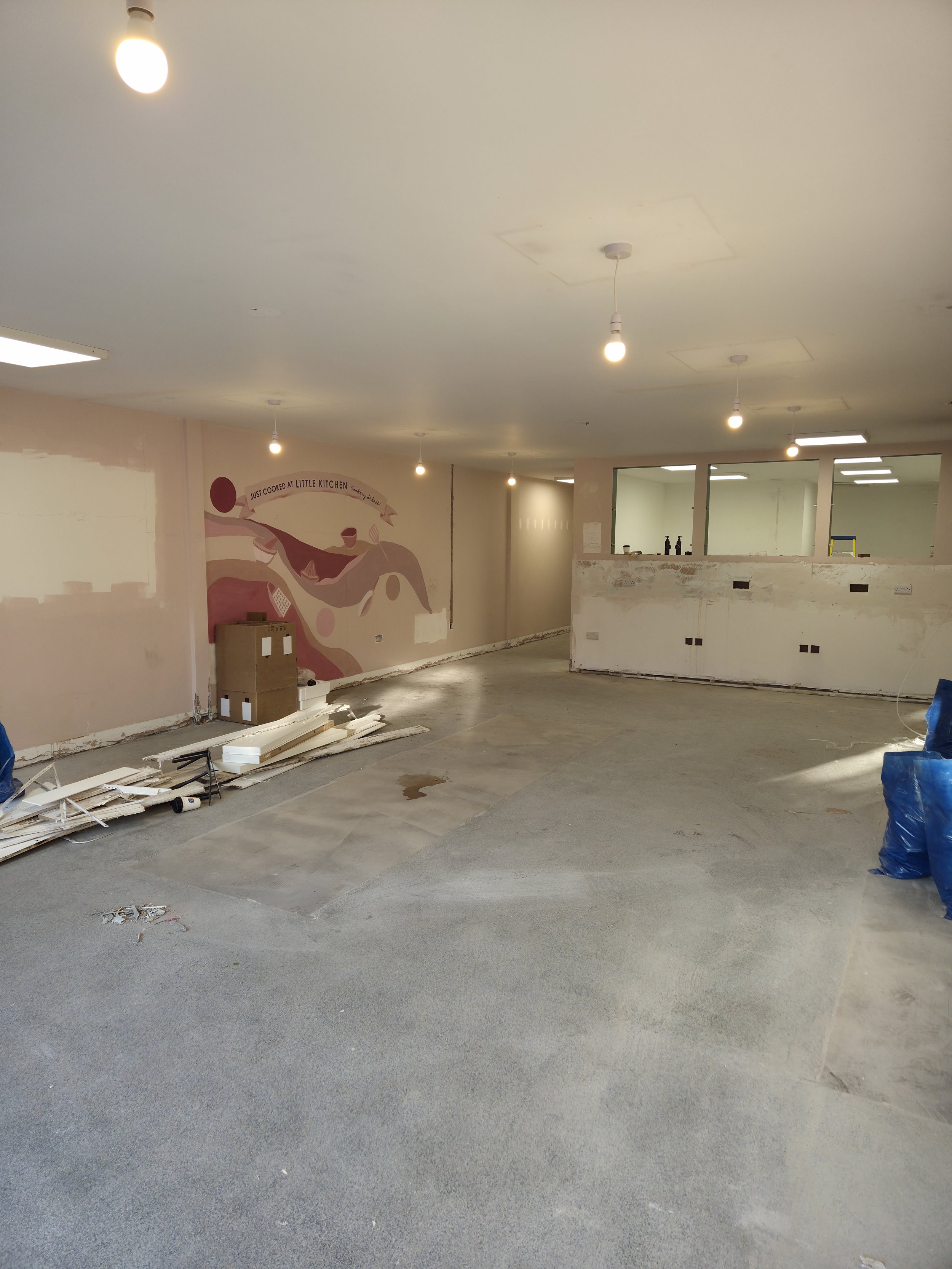 Interior of a room under construction with unfinished walls, hanging light bulbs, and construction materials on the floor. A mural with abstract shapes and the phrase 'Just cooked at Little Kitchen' is painted on one wall.