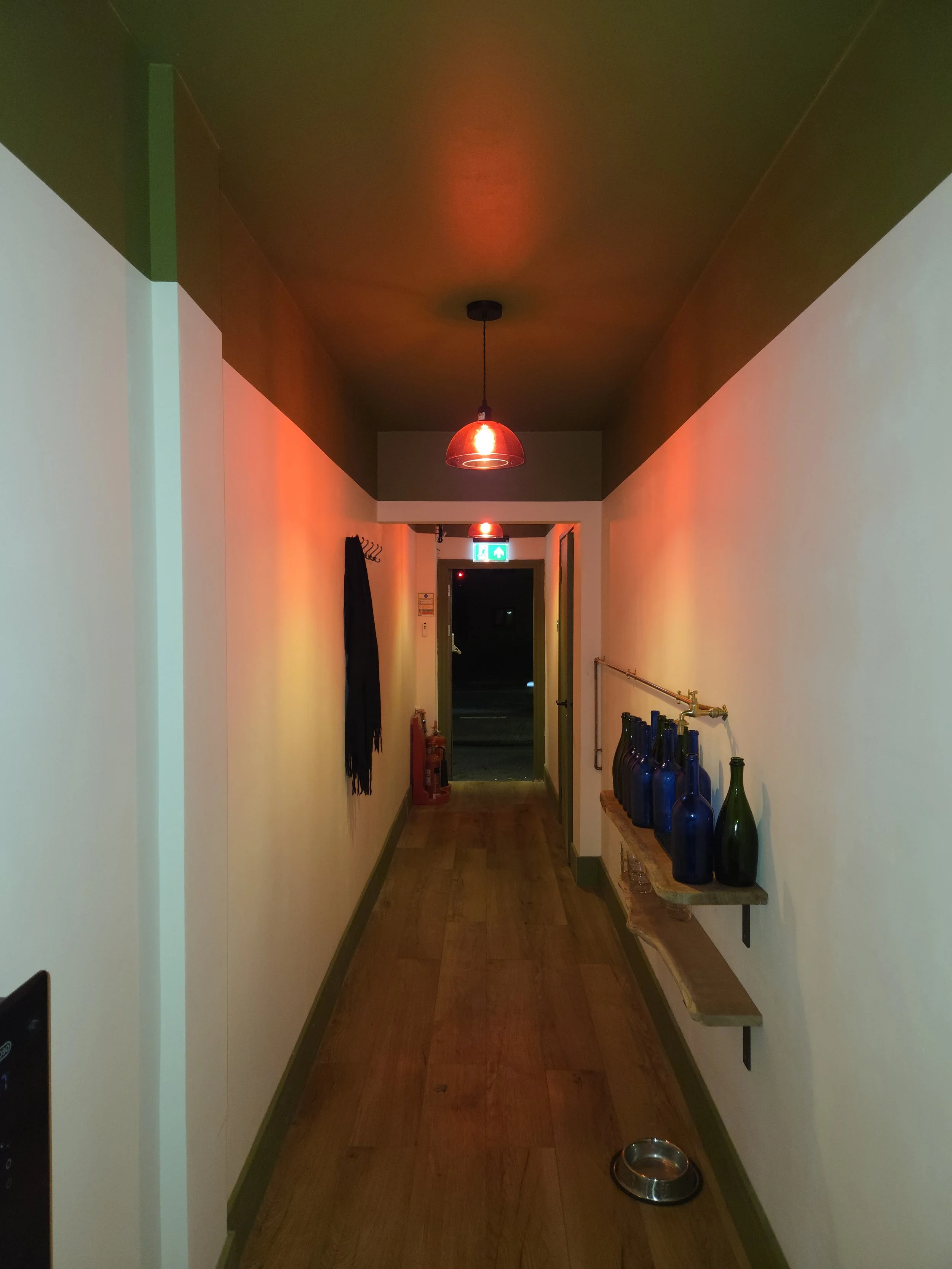 A narrow hallway with hardwood flooring and white walls illuminated by red pendant lights, featuring a coat hook with a dark scarf, a row of blue and green bottles on a shelf, a fire extinguisher, an exit sign, and a door at the end.