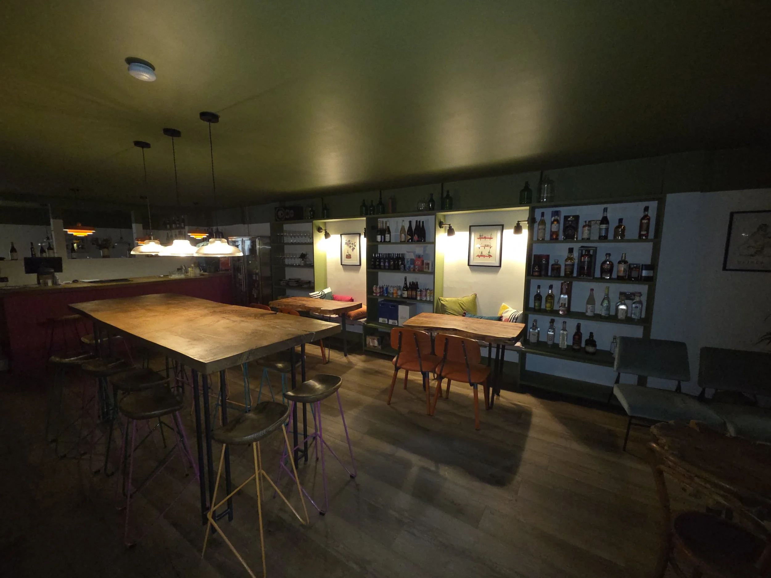Dimly lit restaurant interior with wooden tables and chairs, shelving with bottles on the wall, and warm lighting.