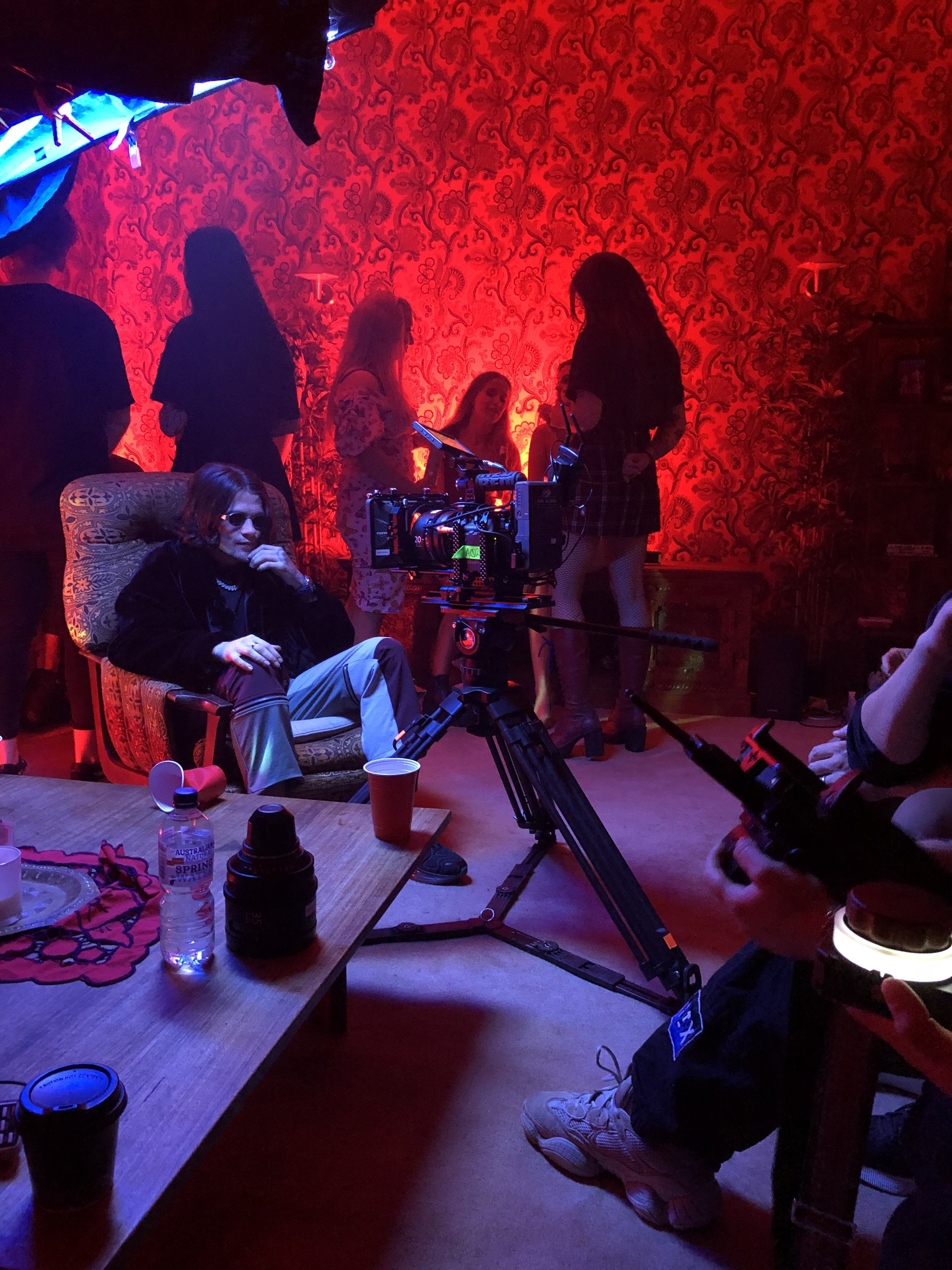 Filming scene in a dimly lit room with red lighting, featuring a person sitting in an armchair and a camera on a tripod nearby. Several people are standing in the background against a patterned red wall. A table with drinks is in the foreground.