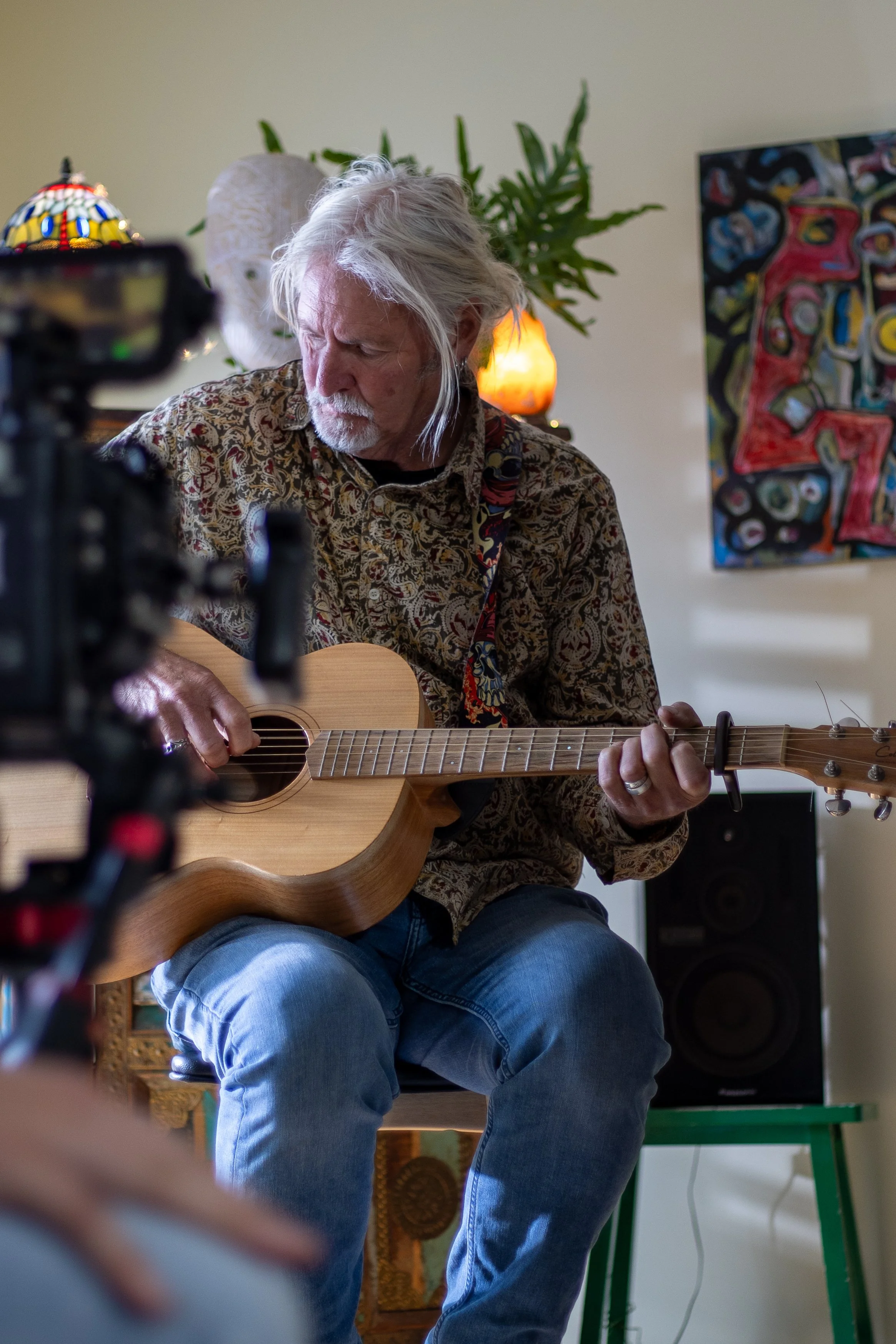 An elderly man with white hair and a beard playing an acoustic guitar indoors, sitting on a chair with a colorful art piece on the wall behind him, a lamp, a potted plant, and a camera filming him.