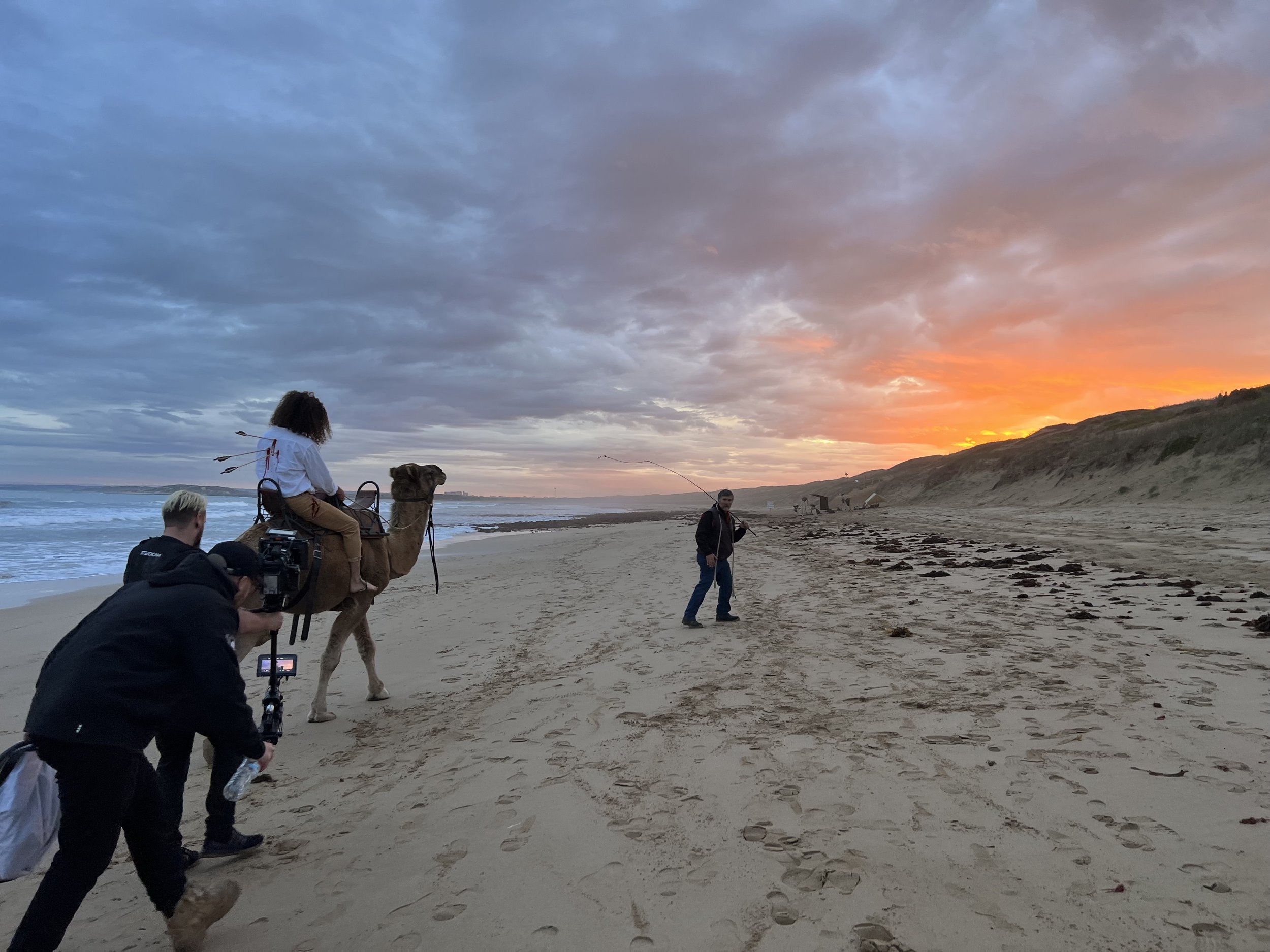 Person riding a camel on a beach during sunset, accompanied by a film crew with cameras, capturing the scene. A person in front is guiding the camel. The sky is vibrant with orange and purple hues.
