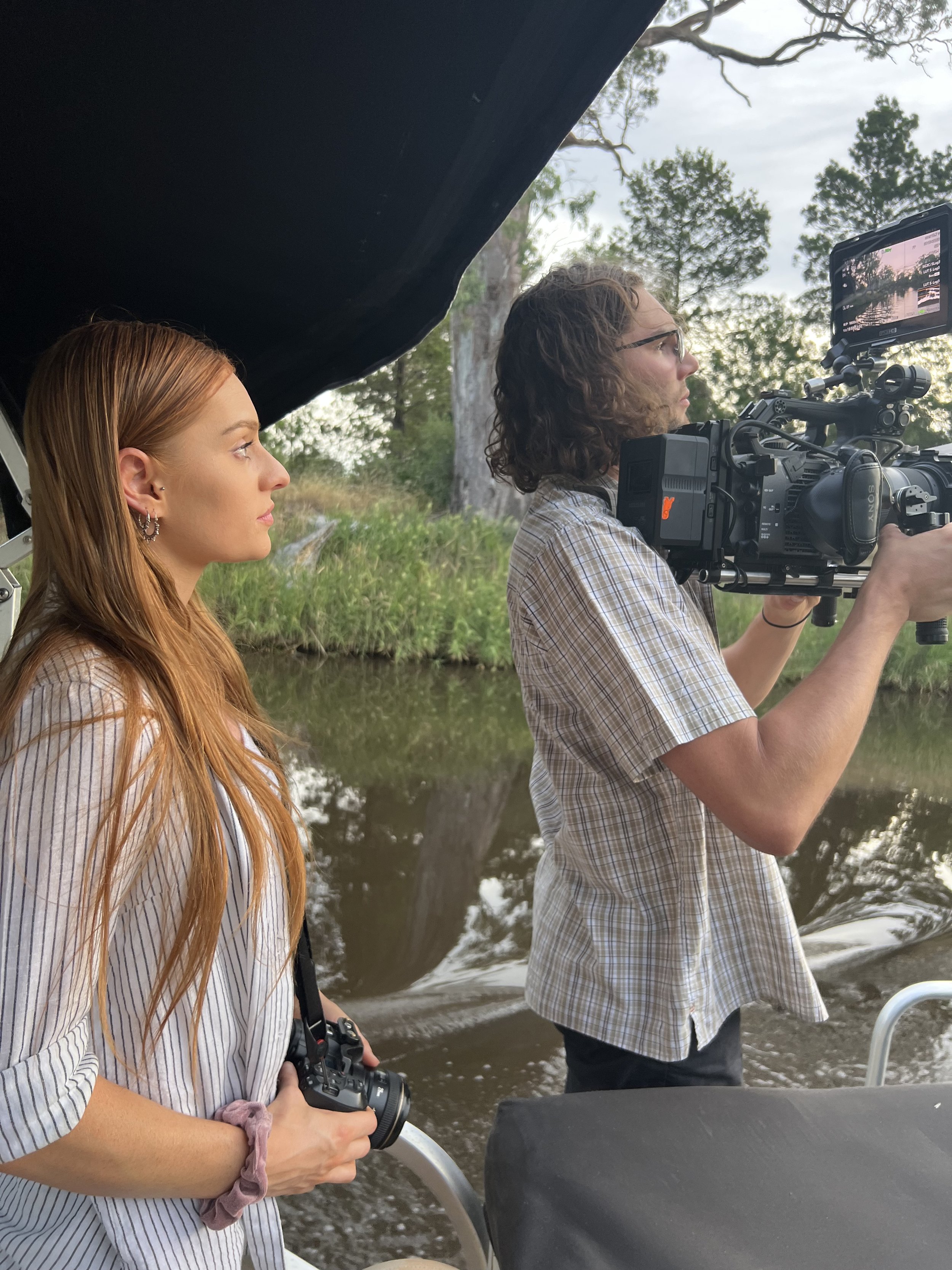 Two individuals on a boat; one woman holding a camera, and one man operating a video camera. They appear to be filming or photographing a scenic area with trees and water in the background.
