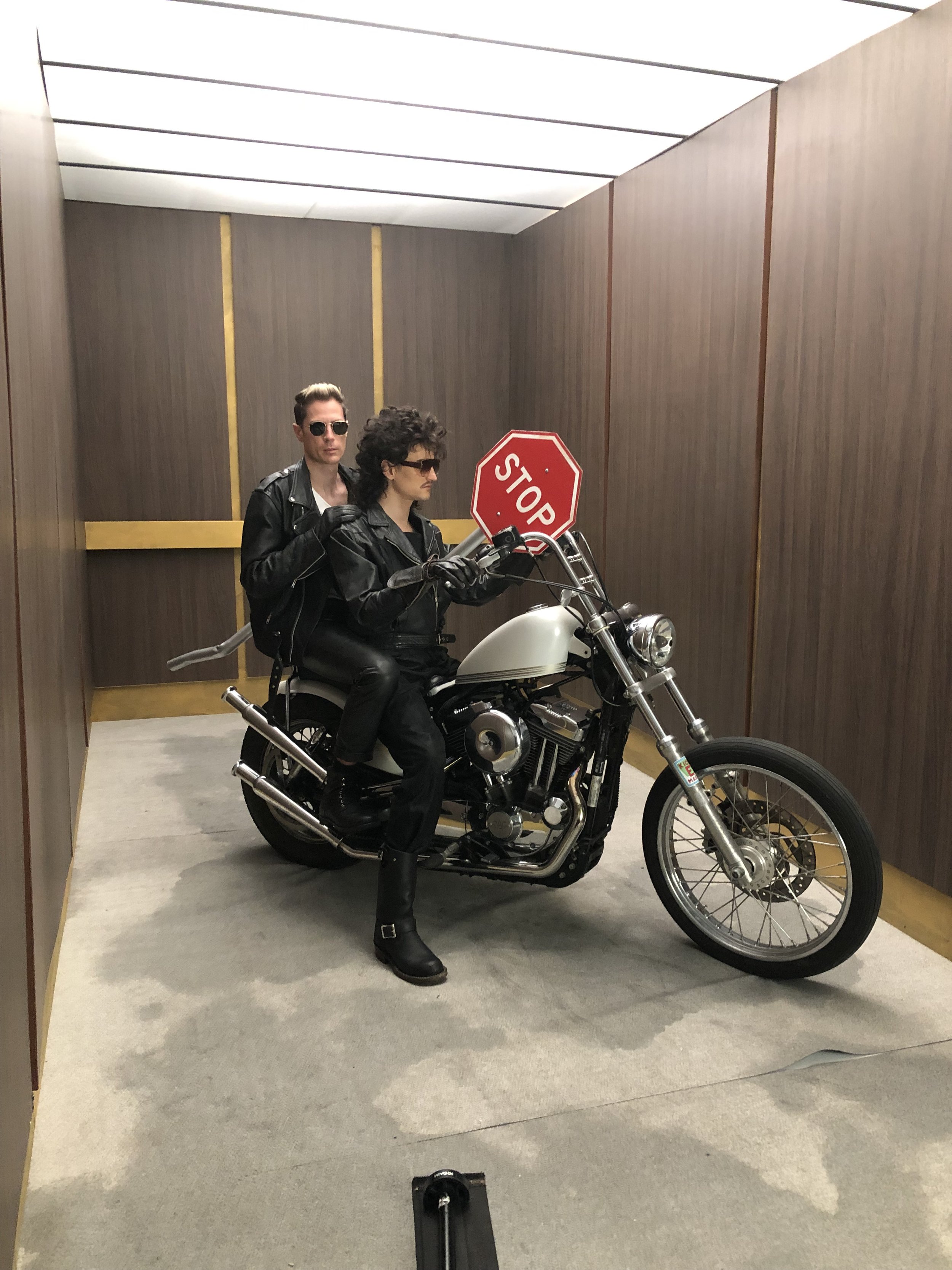 Two people in leather jackets on a motorcycle in an elevator with a stop sign attached to the handlebar.