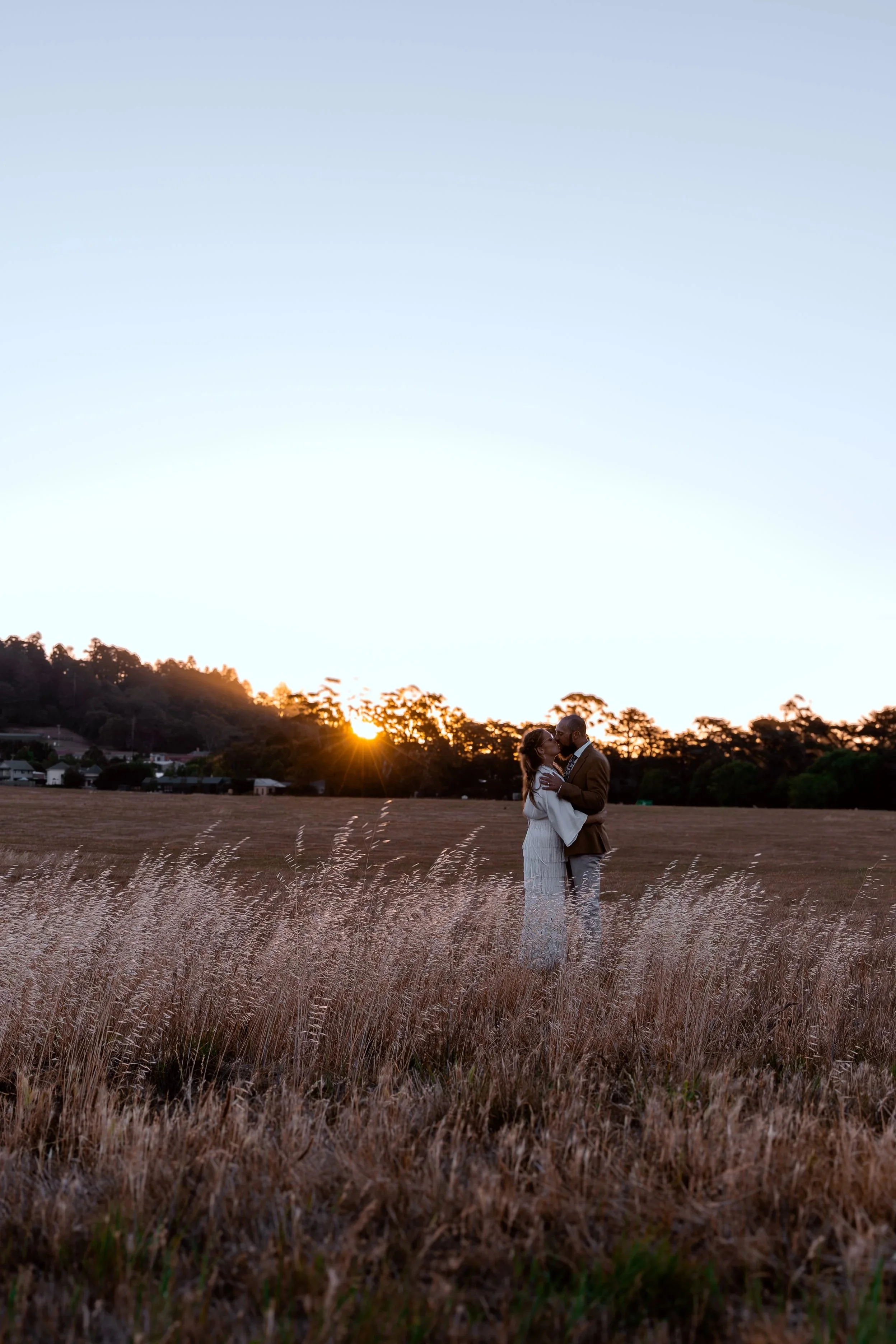 A couple stands close together embracing in a field during sunset, with trees and houses in the background.