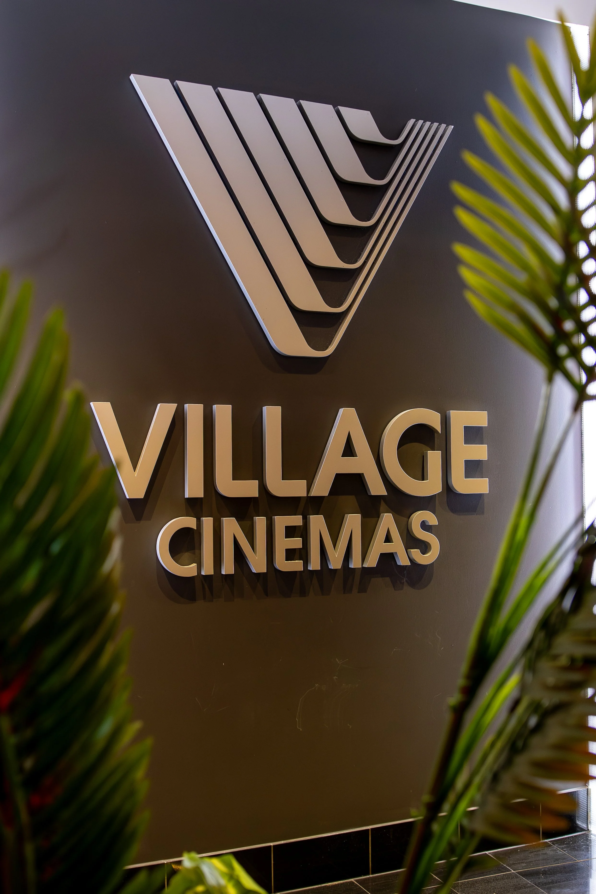 Sign for Village Cinemas seen through green plants, mounted on a dark wall.