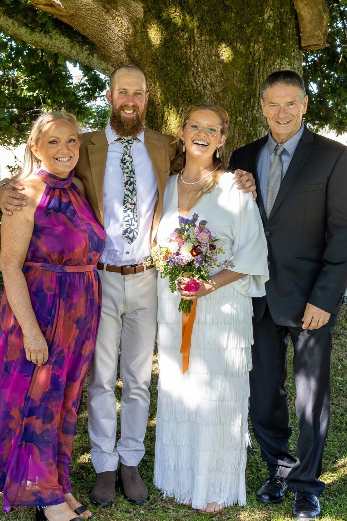 A group of five people standing outdoors in front of a large tree, smiling and posing for a photo, with a woman holding a bouquet of flowers in the center, wearing a white dress, surrounded by two men and two women in colorful and formal clothing.
