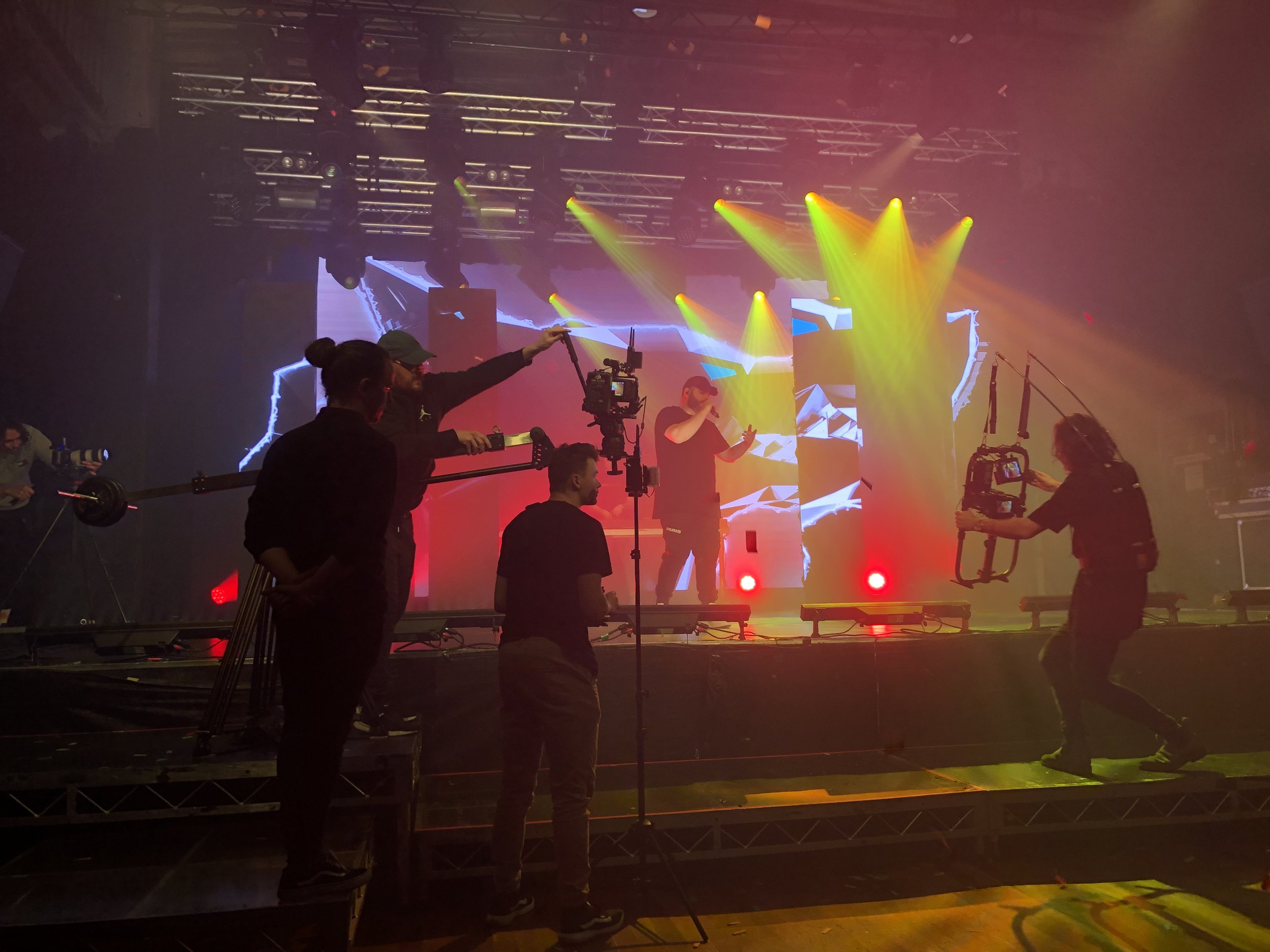 Performers and a camera crew onstage during a live music production with colorful stage lighting and equipment.