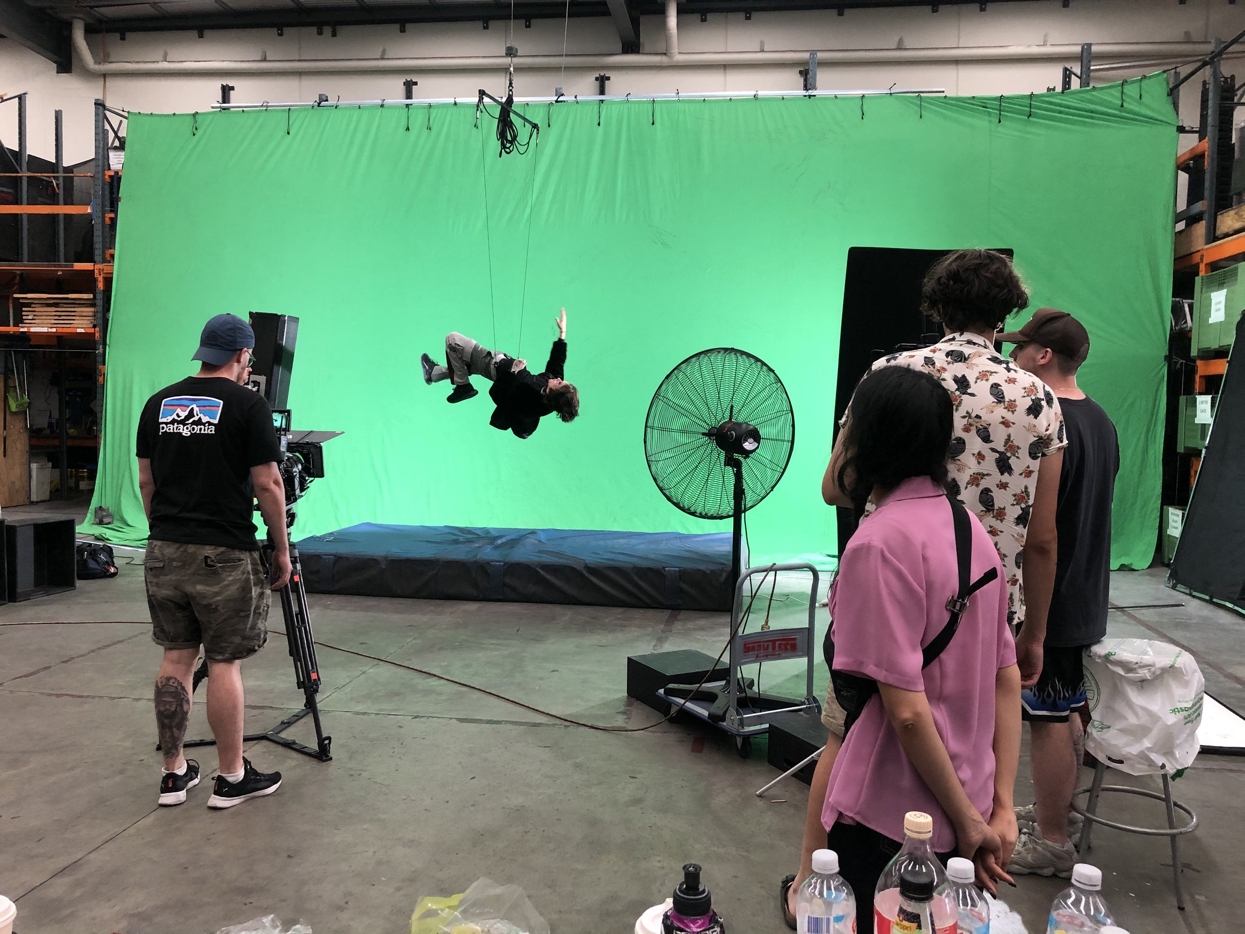 Film crew on set with green screen, actor suspended by wires, camera operator in black t-shirt, equipment and accessories in foreground.
