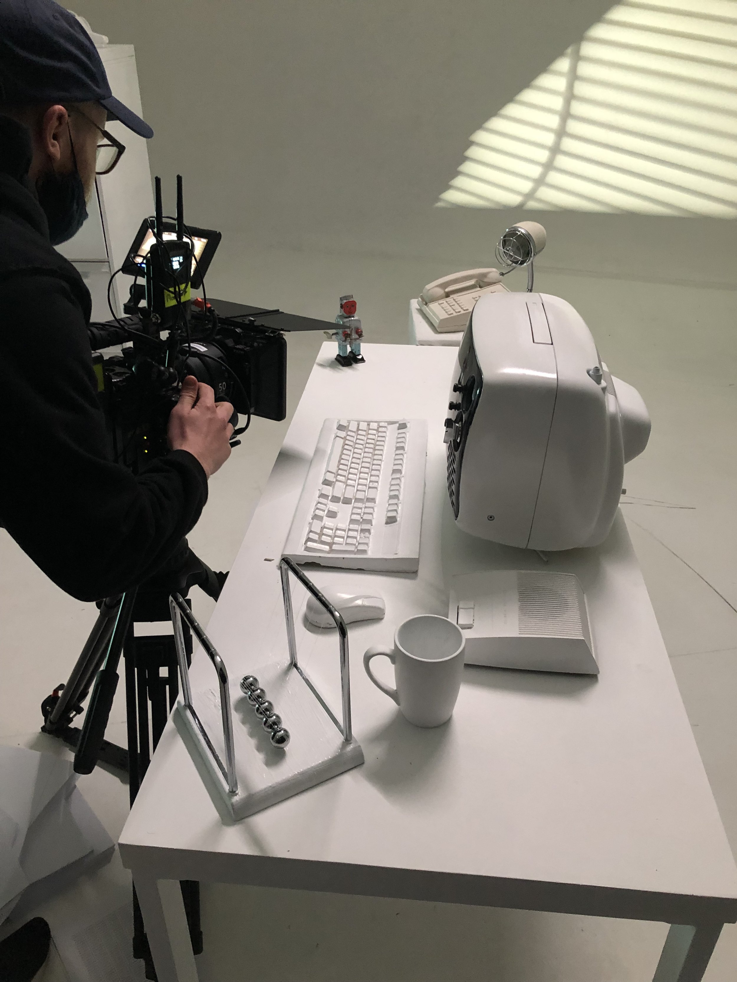 Person setting up a camera to film a white desk with a vintage computer, keyboard, a Newton's cradle, a coffee mug, a toy figure, and a telephone.