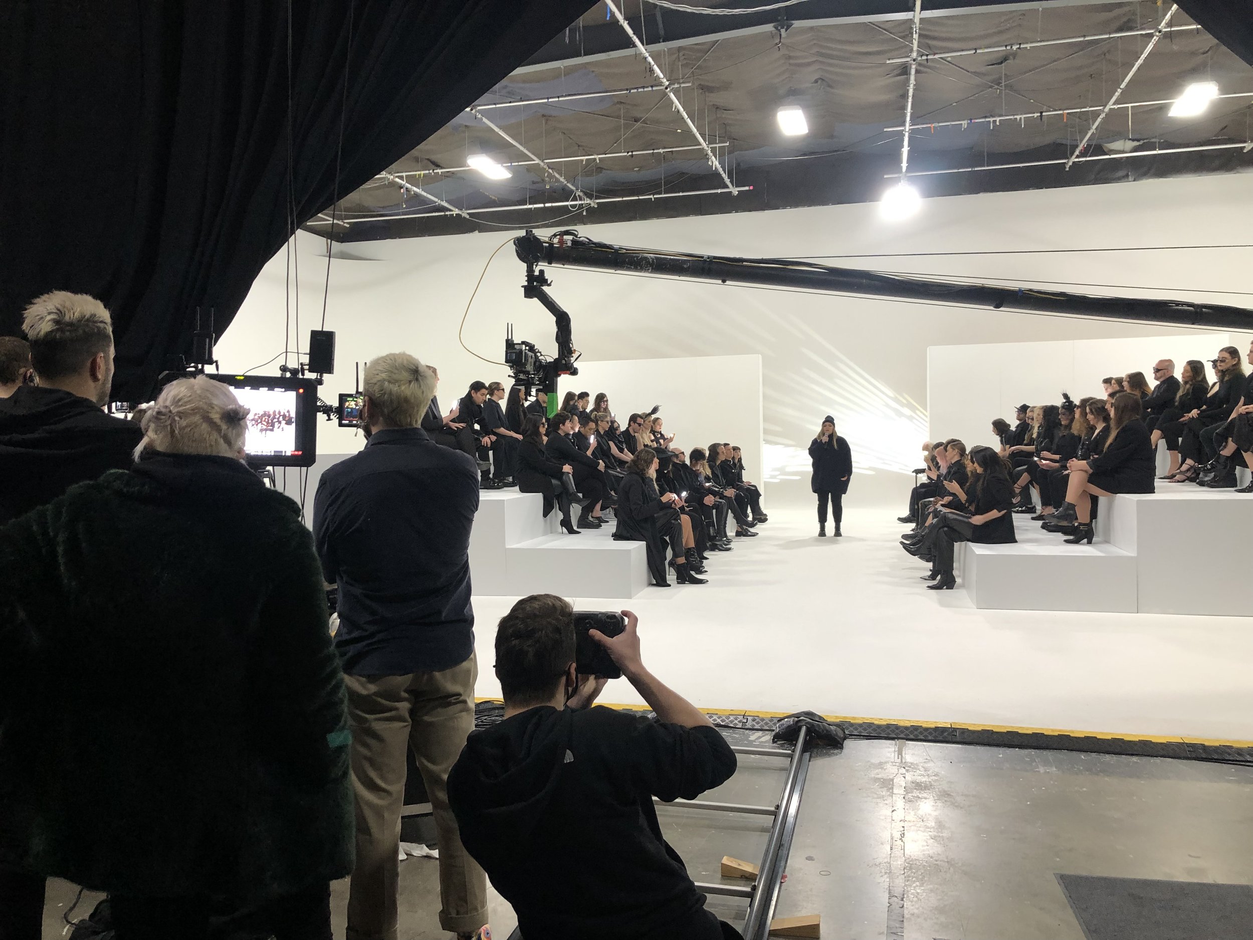 A studio with people seated on tiered platforms, a person speaking, and a film crew with cameras and equipment.