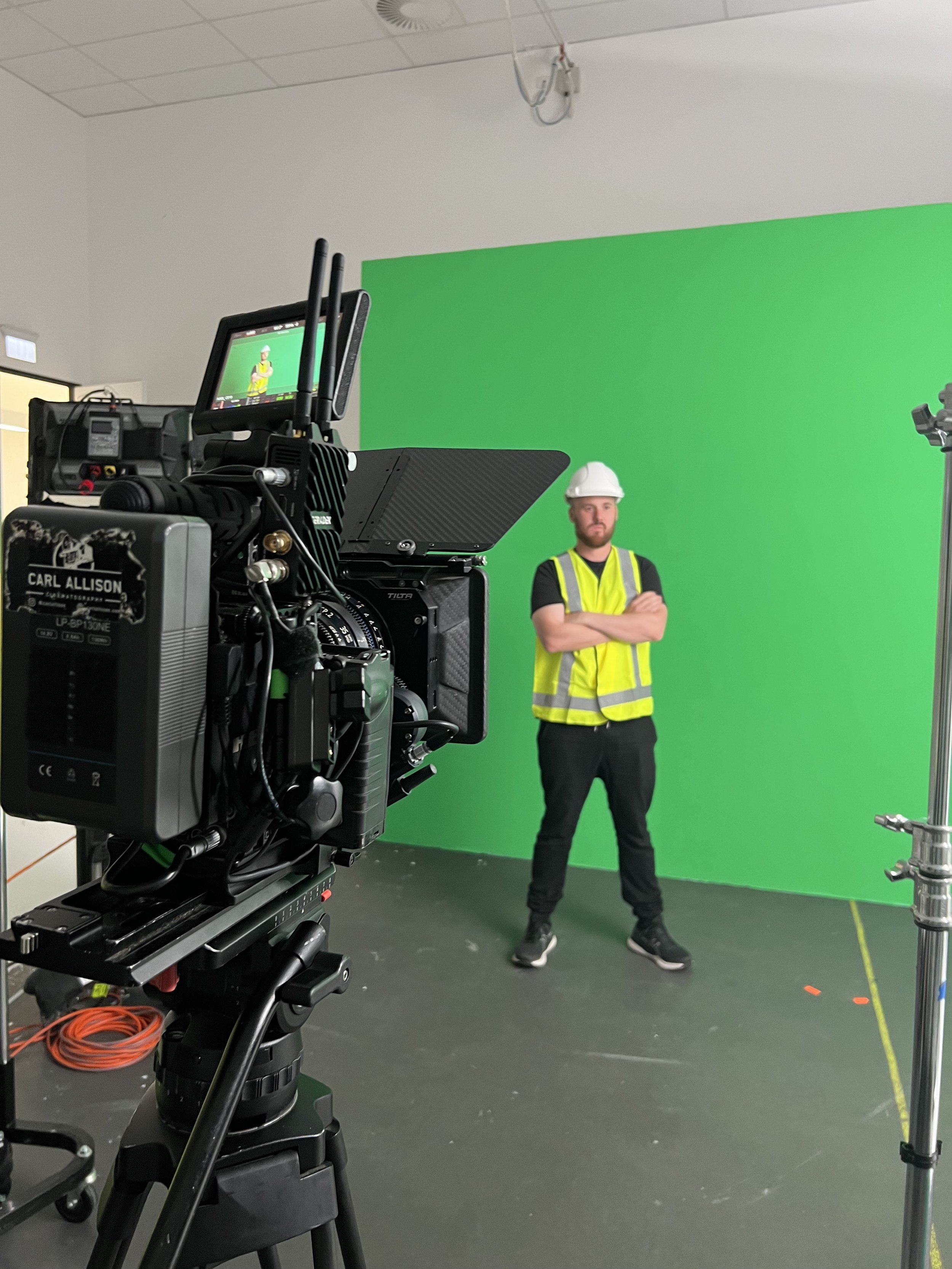 A person wearing a hard hat and high-visibility vest stands in front of a green screen, with a professional video camera setup in the foreground on the left.