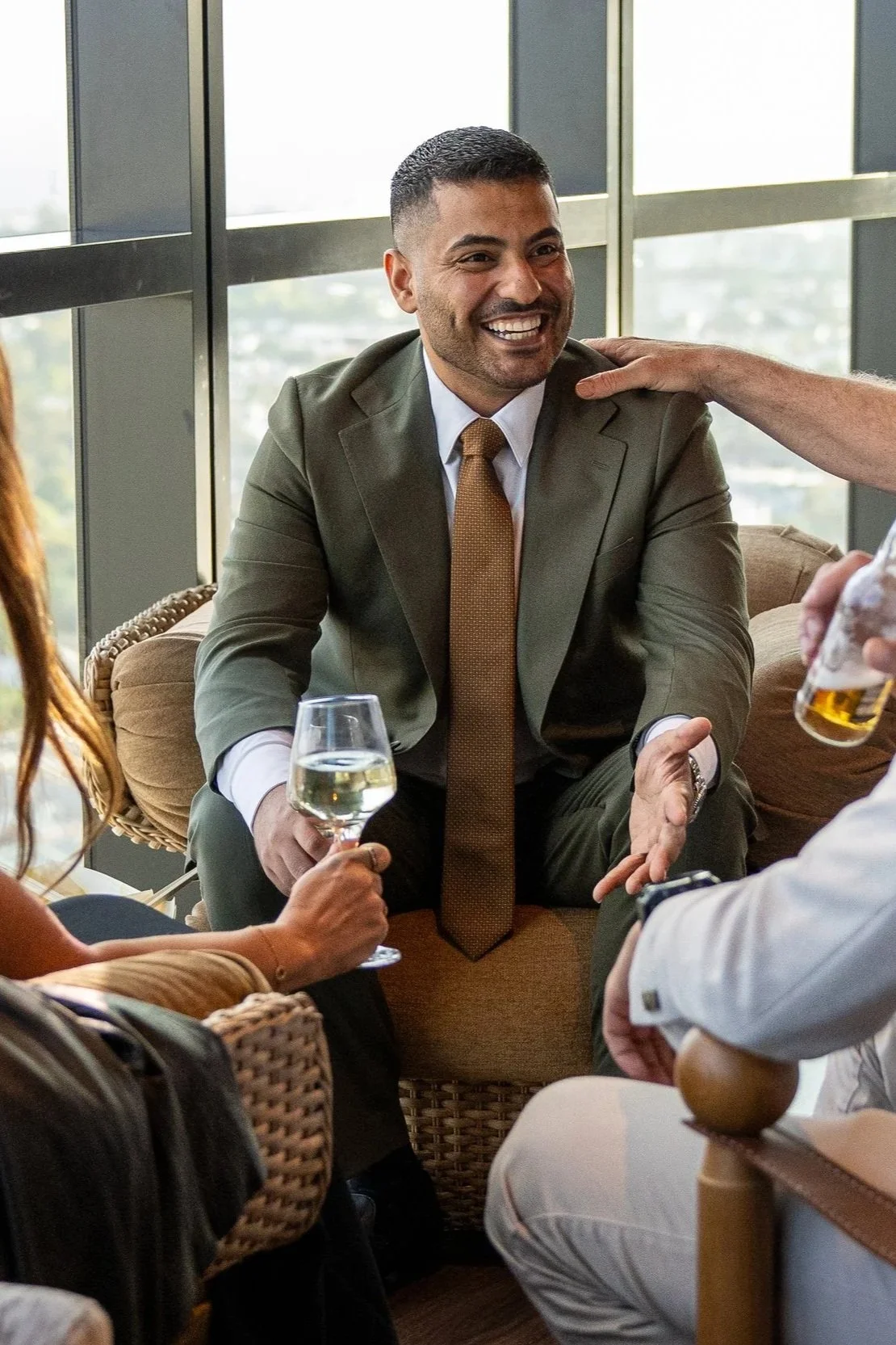 A man in a green suit, white shirt, and brown tie is smiling while sitting on a sofa, holding a glass of white wine. He is surrounded by people, some of whom are pouring drinks and engaging in conversation, in a bright room with large windows.