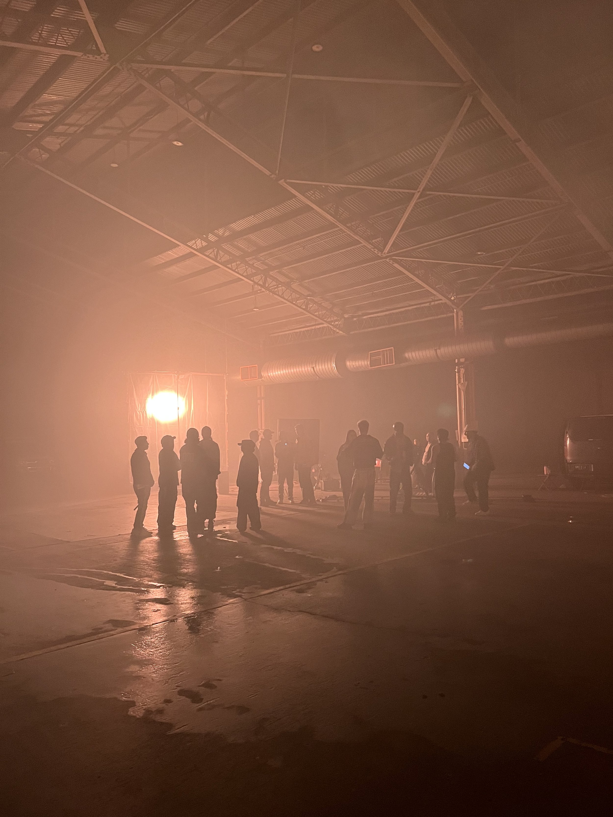 Silhouettes of people gathered in a dimly lit warehouse with a bright light source in the background.