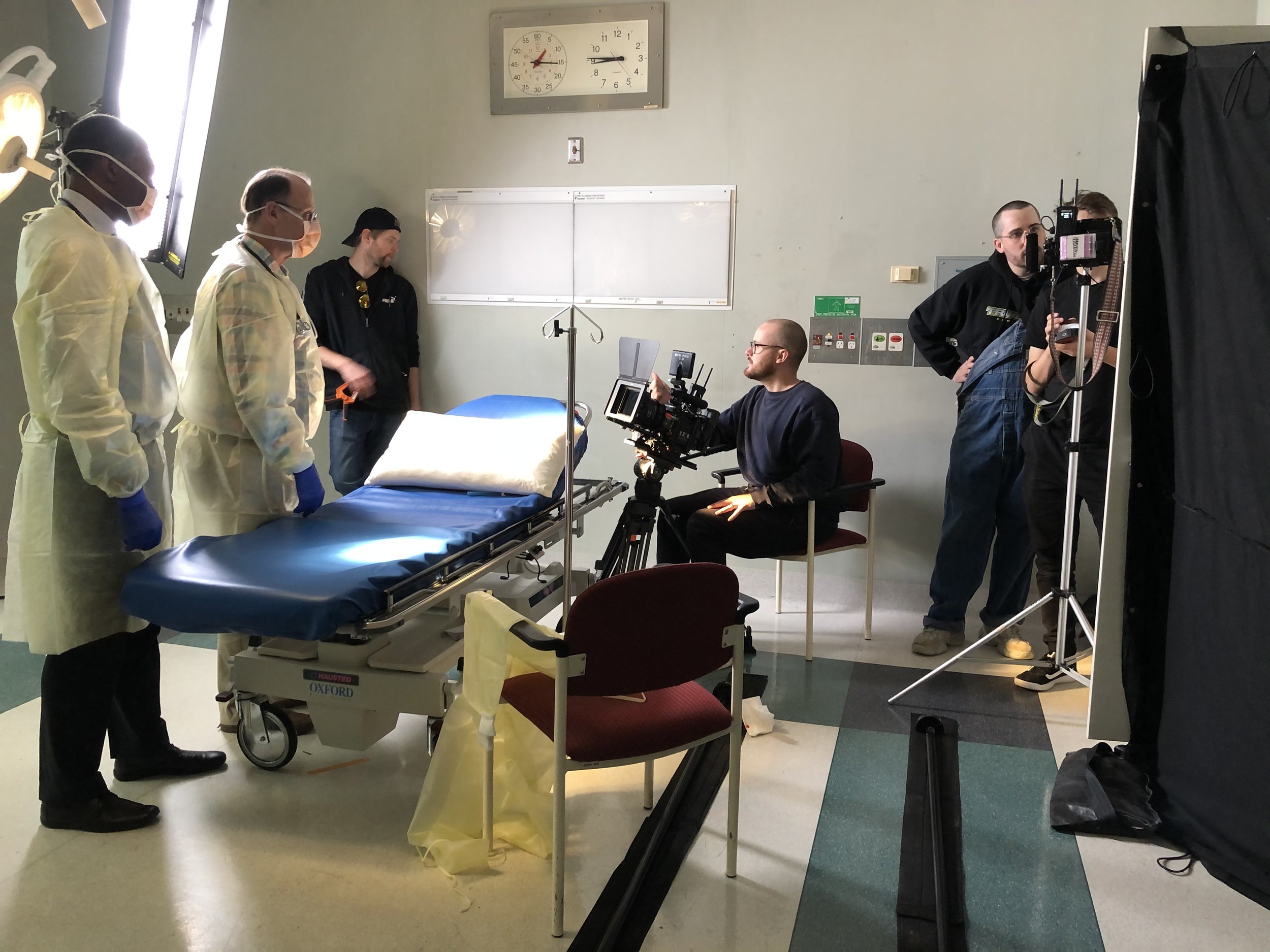 Film crew setting up a scene in a medical setting with actors in protective gear and a gurney.