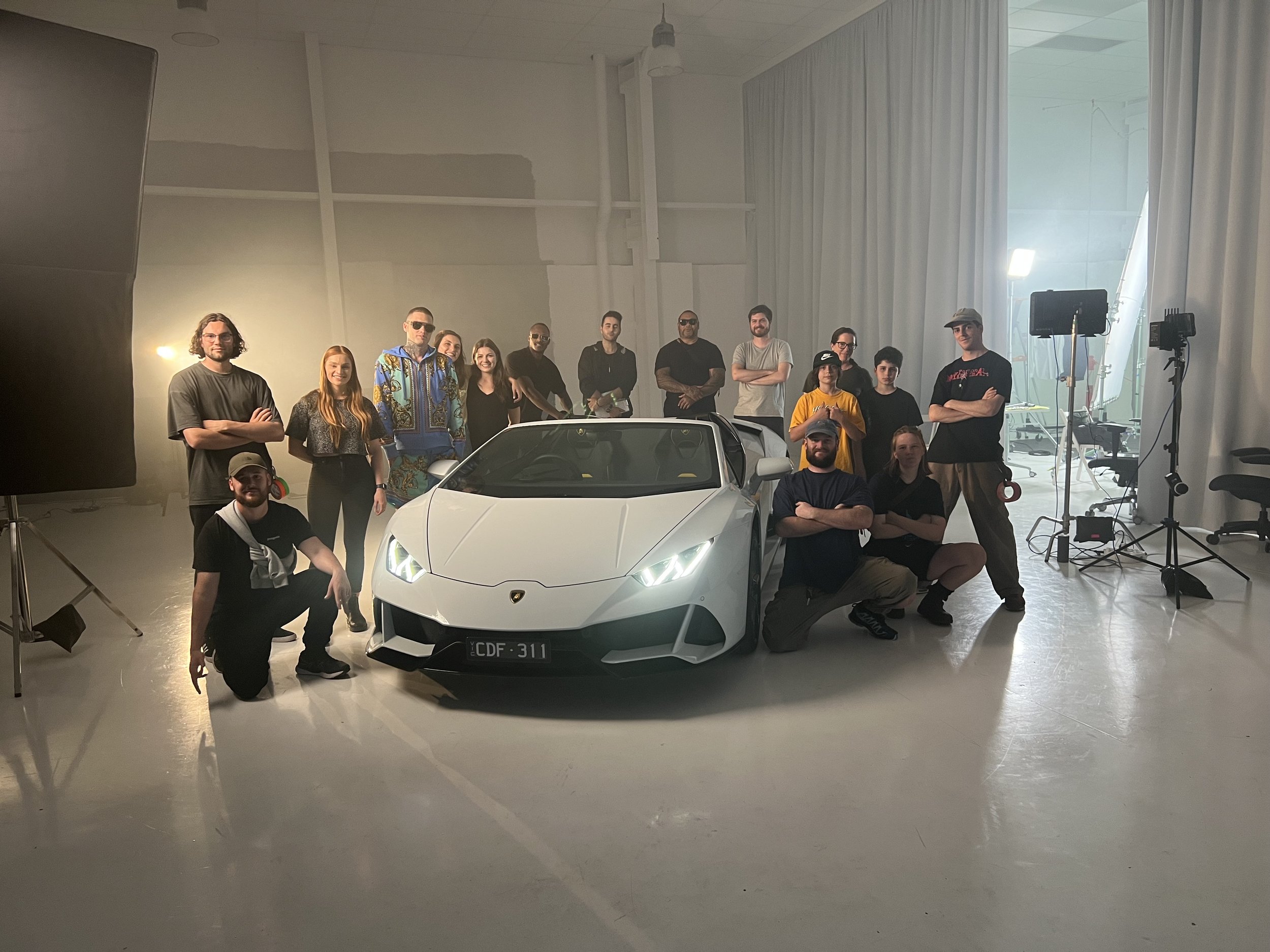 Group of people posed around a white sports car in a studio setting with lighting equipment.