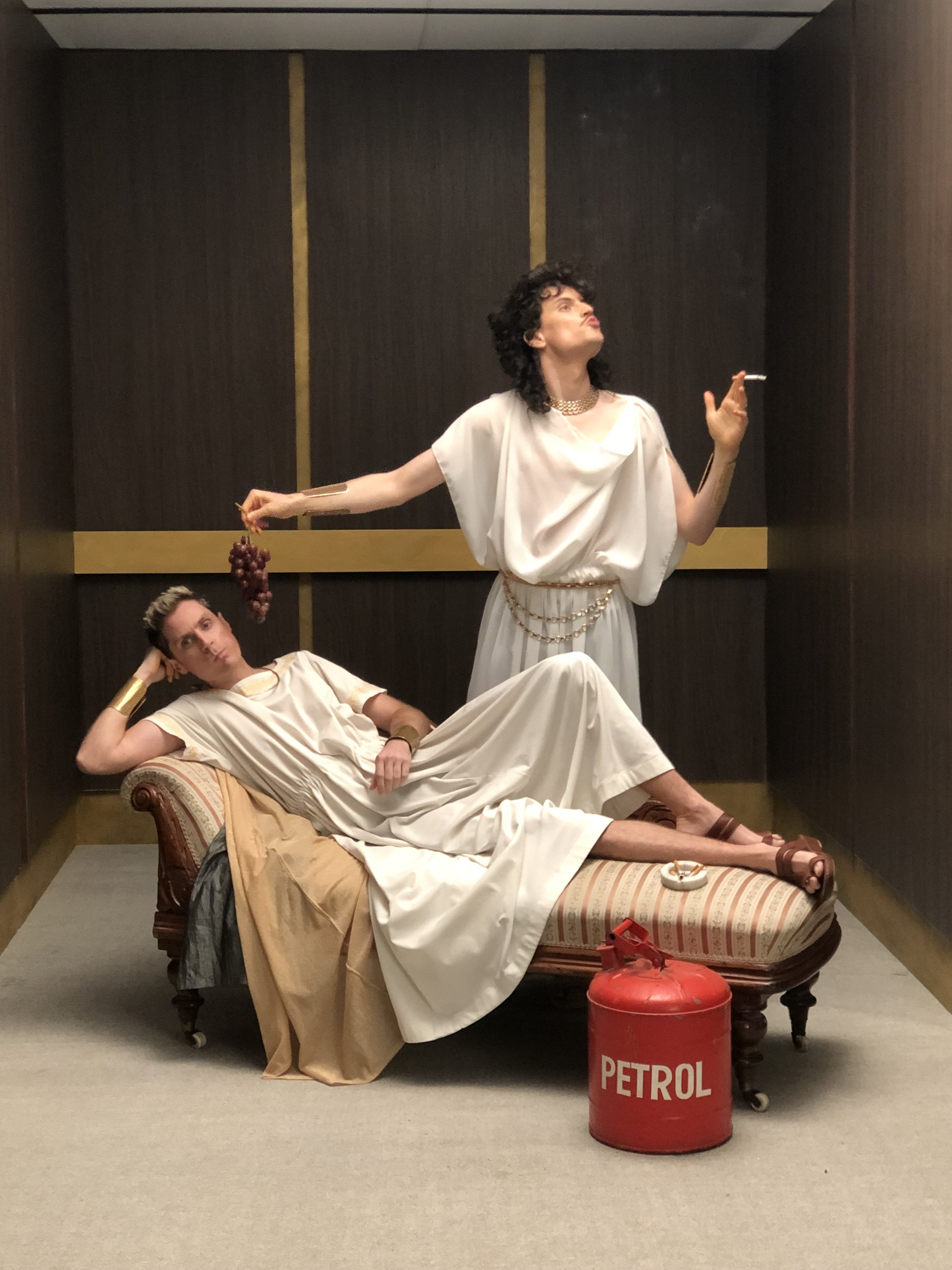 Two people in ancient Roman-style attire, one reclining on a chaise lounge holding grapes, the other standing with a cigarette. A red petrol canister is on the floor.