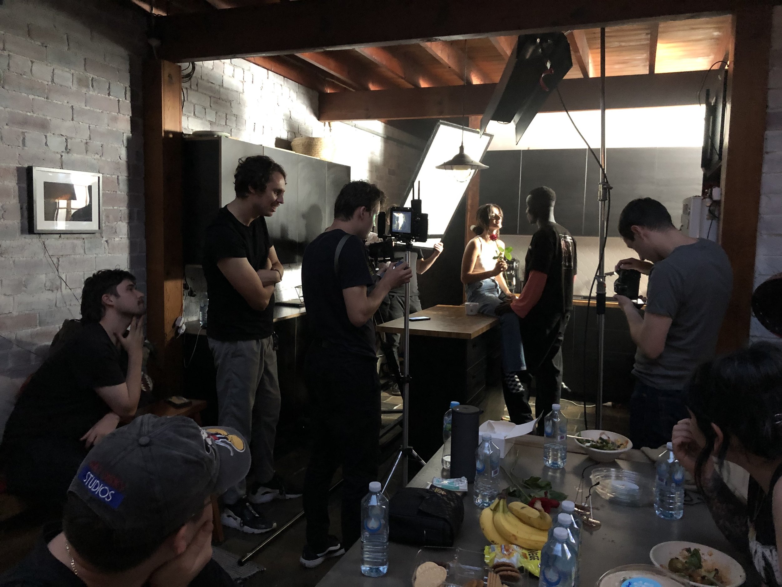 Film crew in a studio setting with cameras and lighting equipment, two actors interacting in the background, table with food and water bottles.