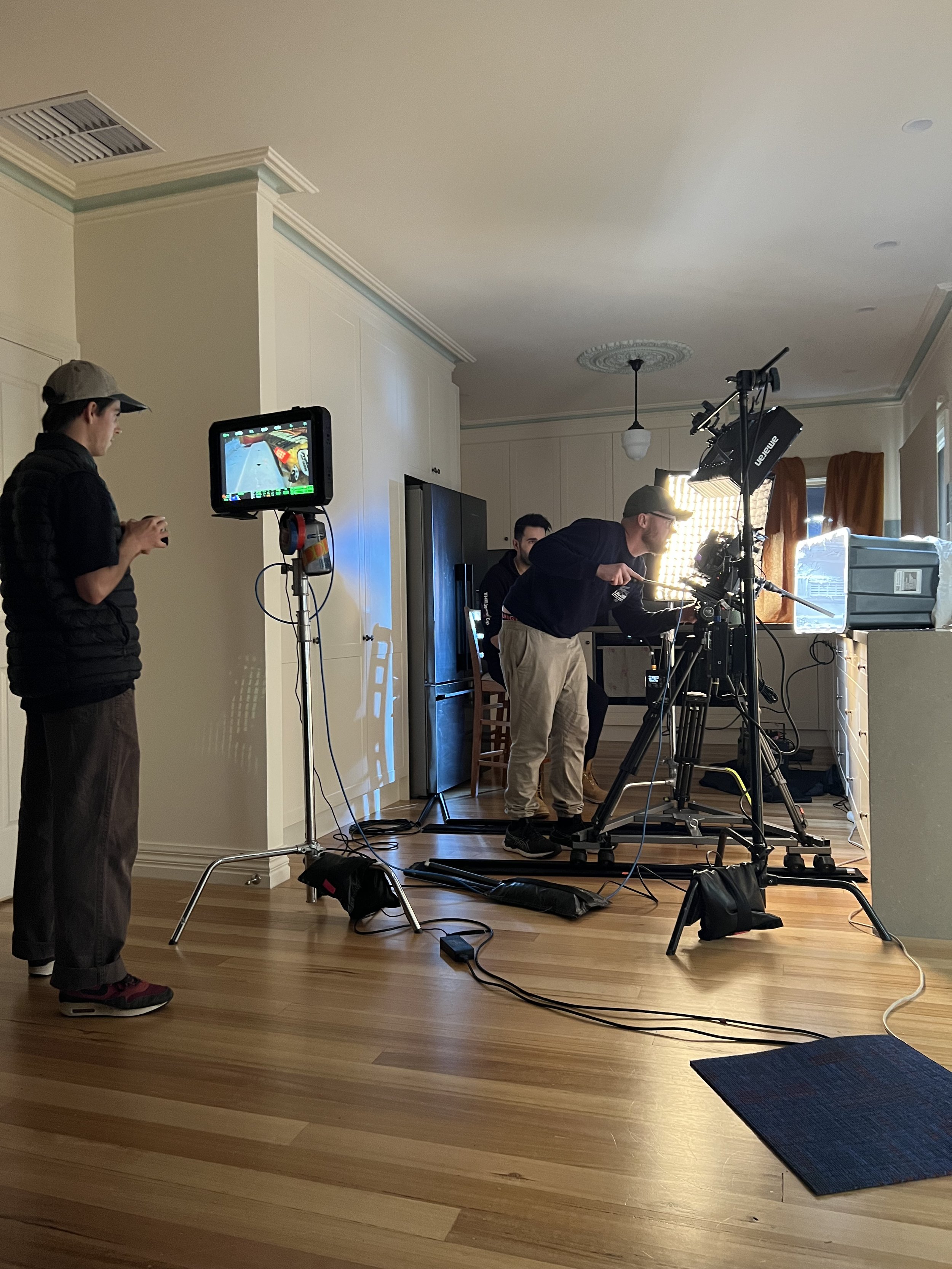 A film crew setting up camera and lighting equipment in a kitchen, with one person holding a monitor.