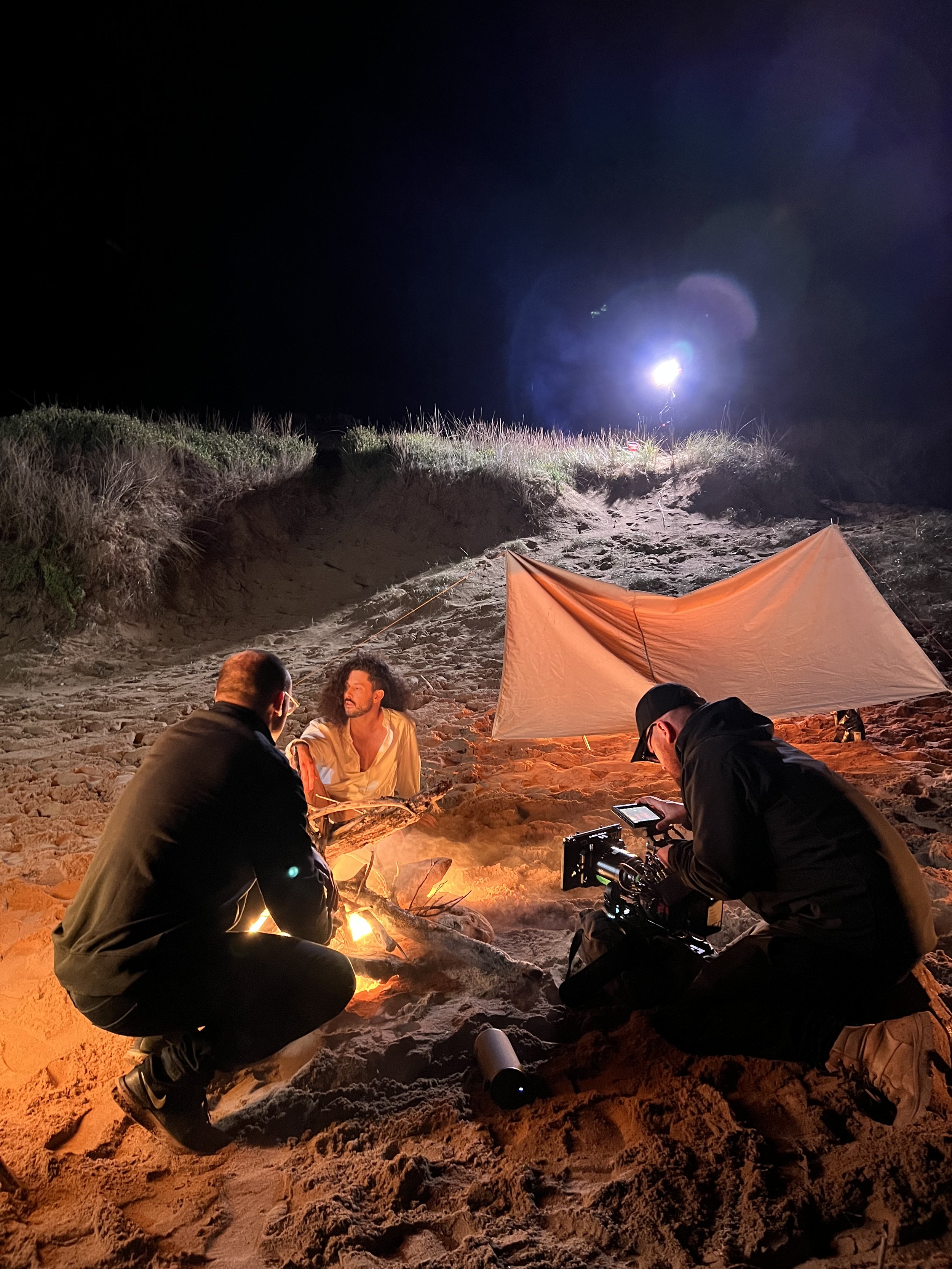 Nighttime film shoot on a sandy beach with a person sitting by a fire, another person filming, and a tarp providing shelter.