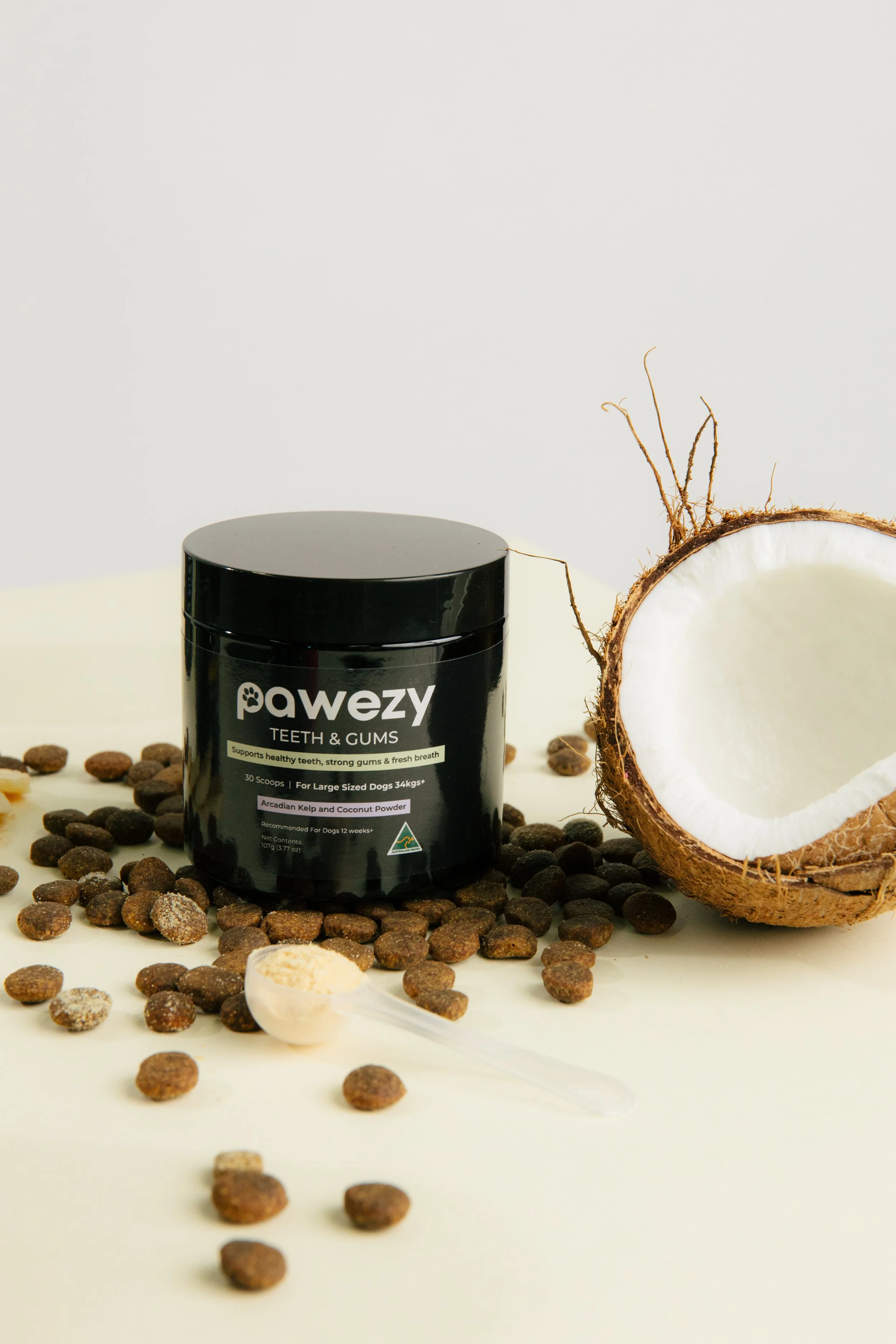 A jar of Pawezy teeth and gums supplement next to a halved coconut and scattered dog treats on a pale yellow surface.