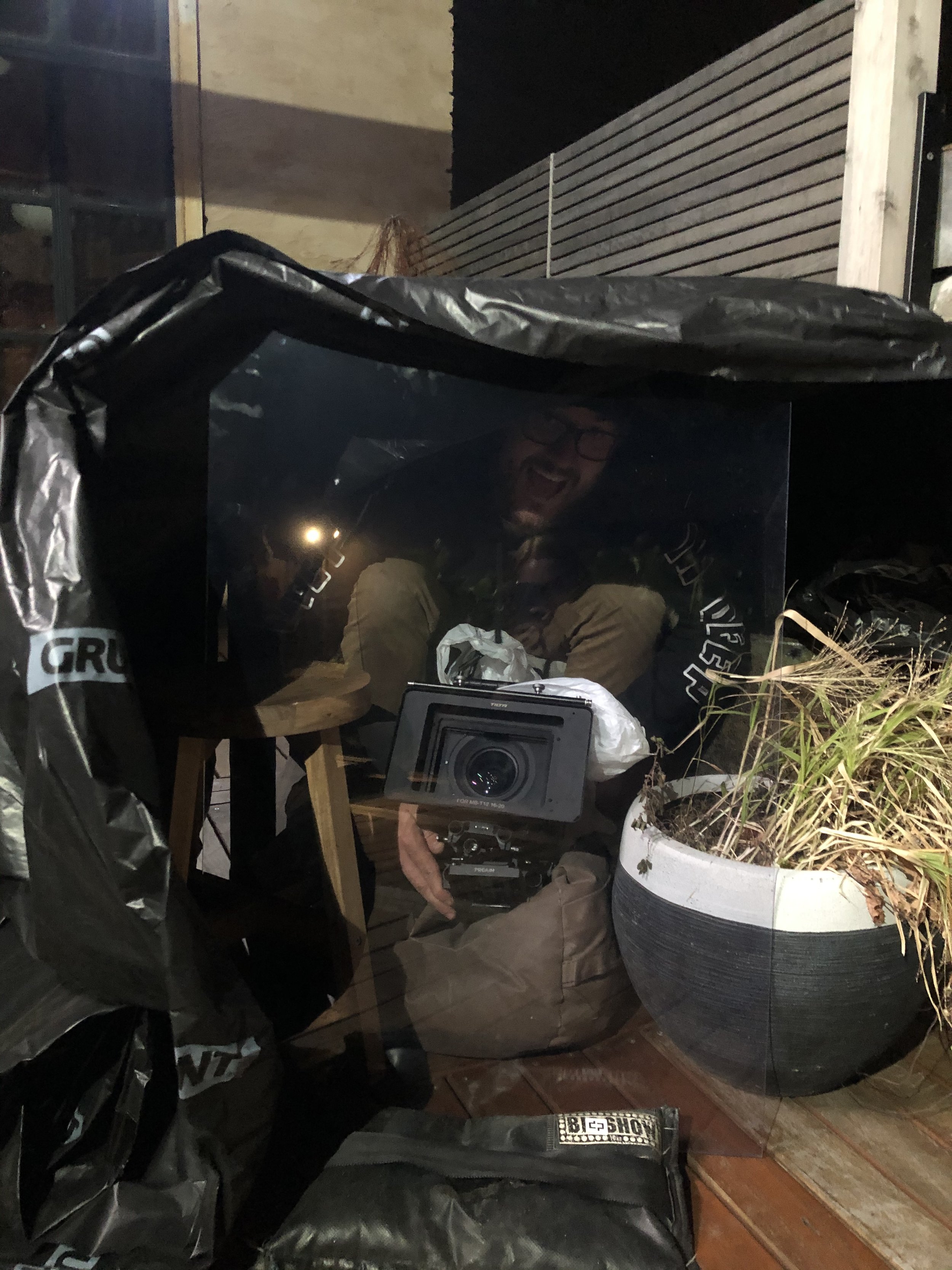 Person inside a makeshift shelter with a camera, surrounded by black plastic sheets and a potted plant on a wooden deck at night.