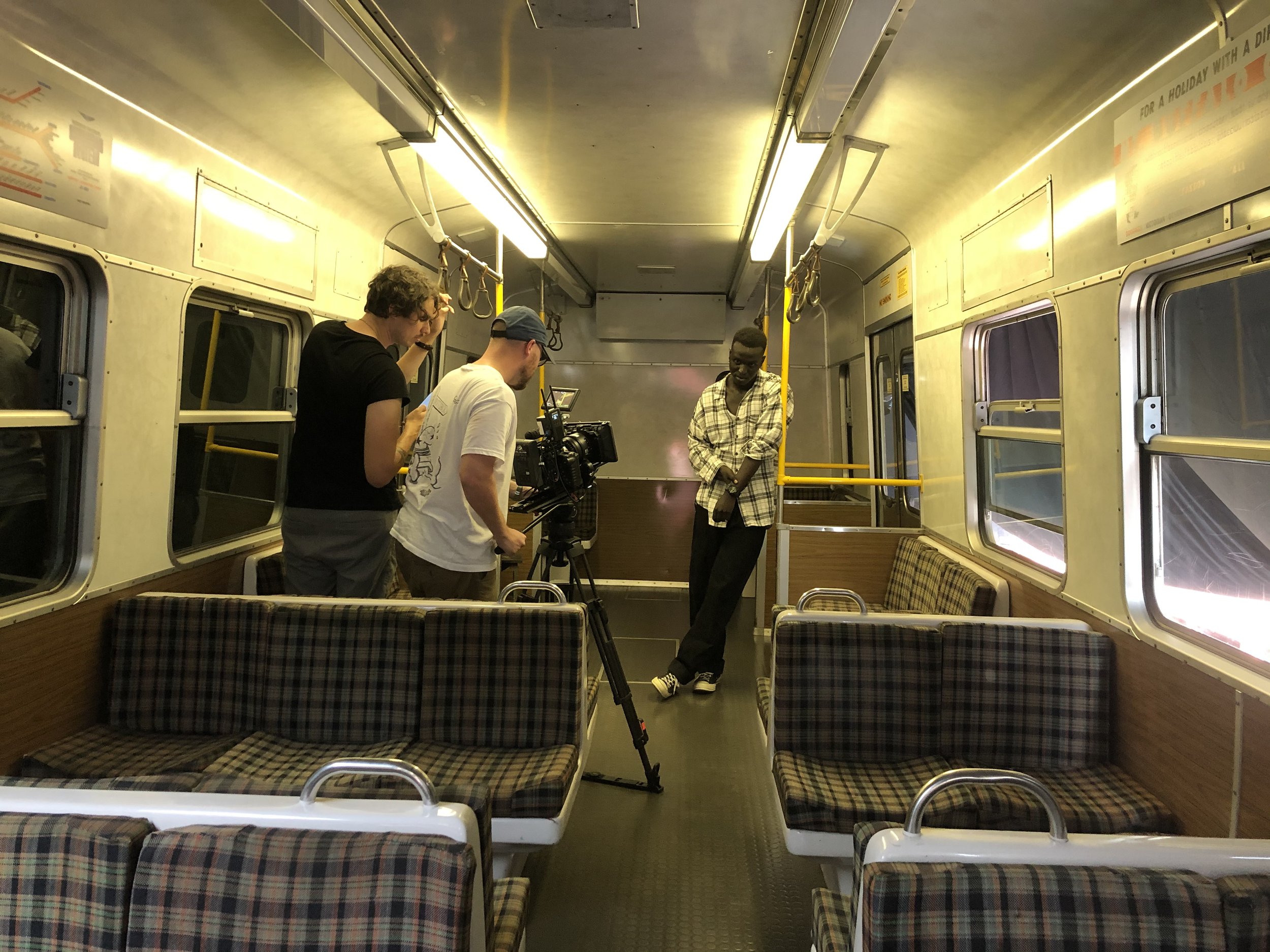 A film crew setting up a camera in an empty subway car with a person standing against a pole.