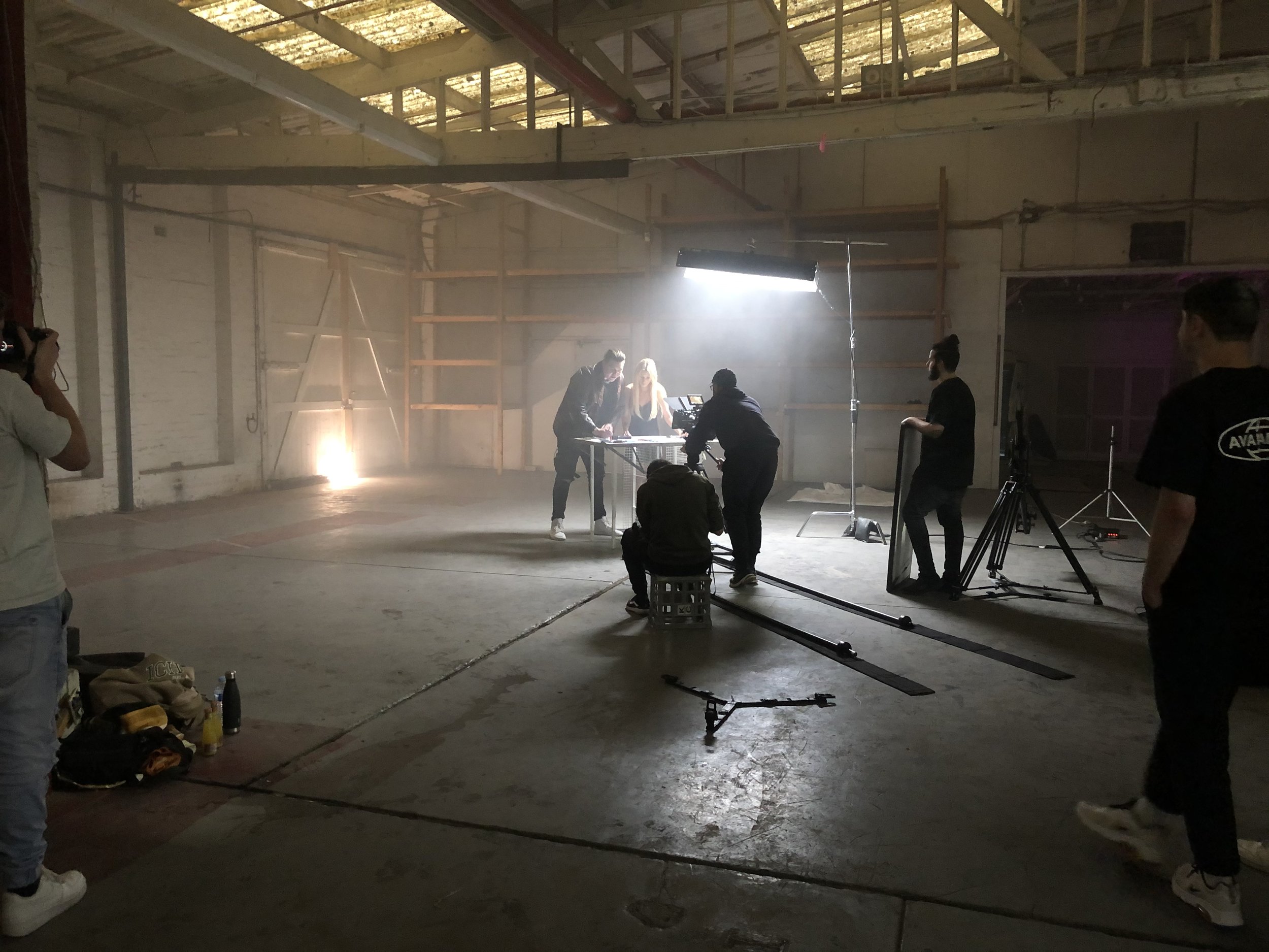 Film crew setting up in an industrial warehouse, with lighting equipment and camera on a track, capturing a scene at a table. People are visible adjusting equipment and directing the setup.