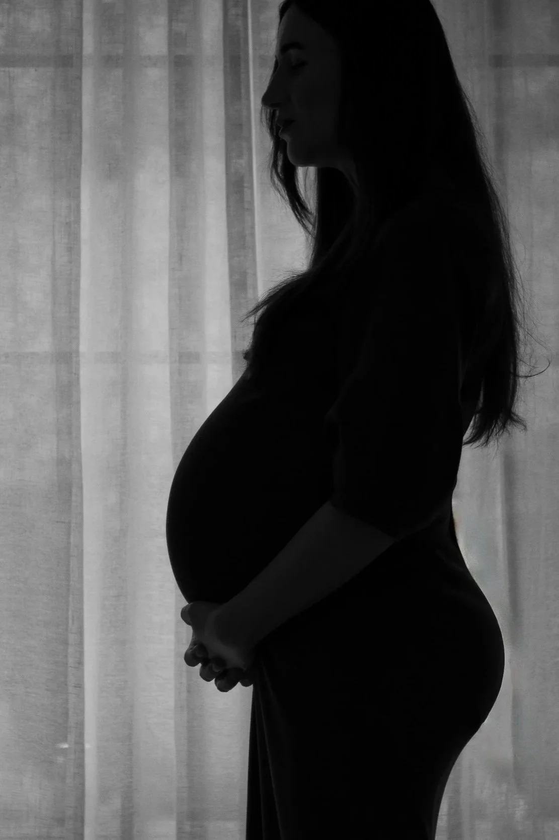 Silhouette of a pregnant woman with long hair, standing sideways with her hands holding her belly, in front of a light-colored curtain.