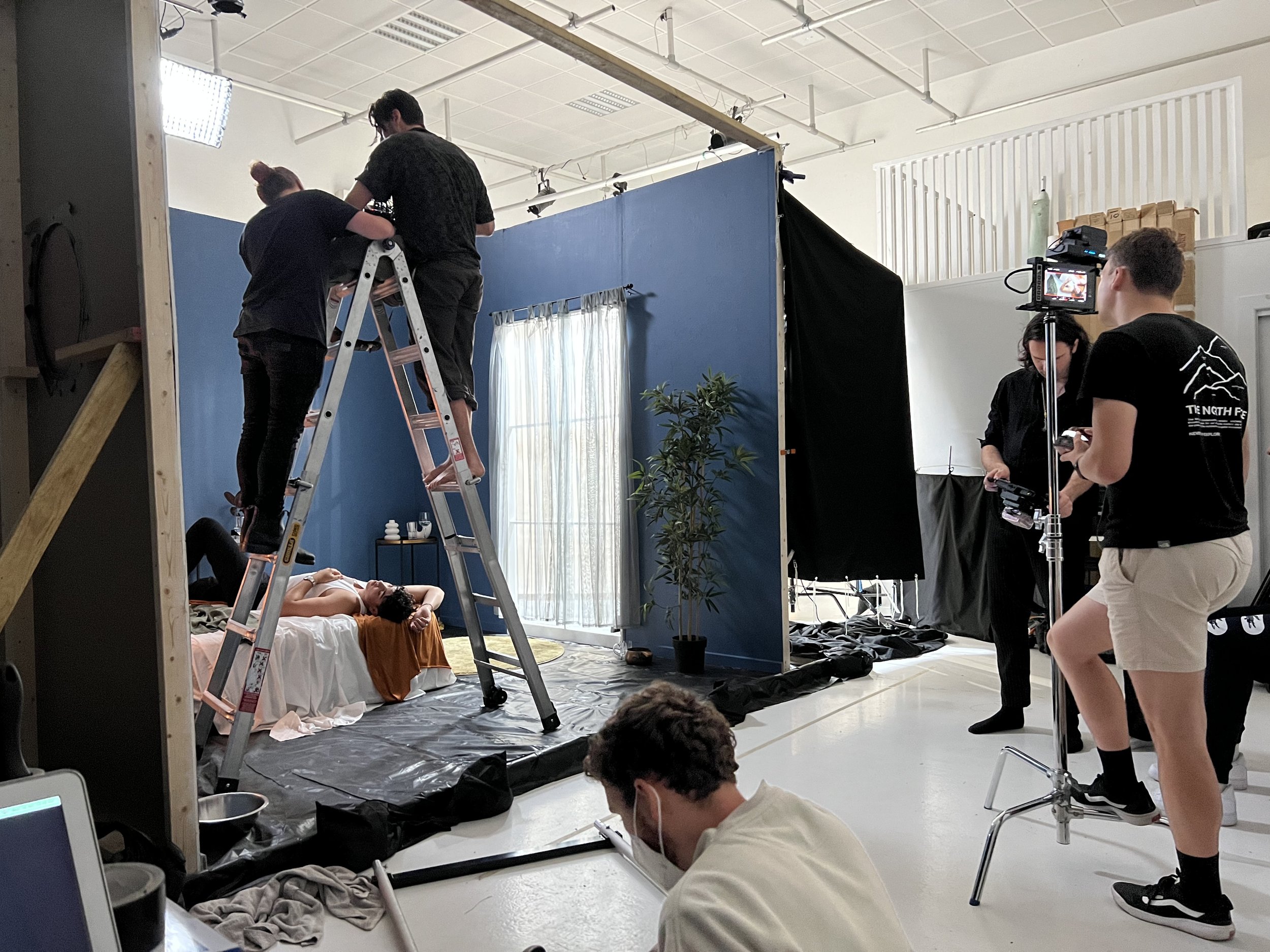 Film crew setting up a scene on a set. A person is lying on a bed with two crew members on a ladder above, and others operating cameras and equipment.