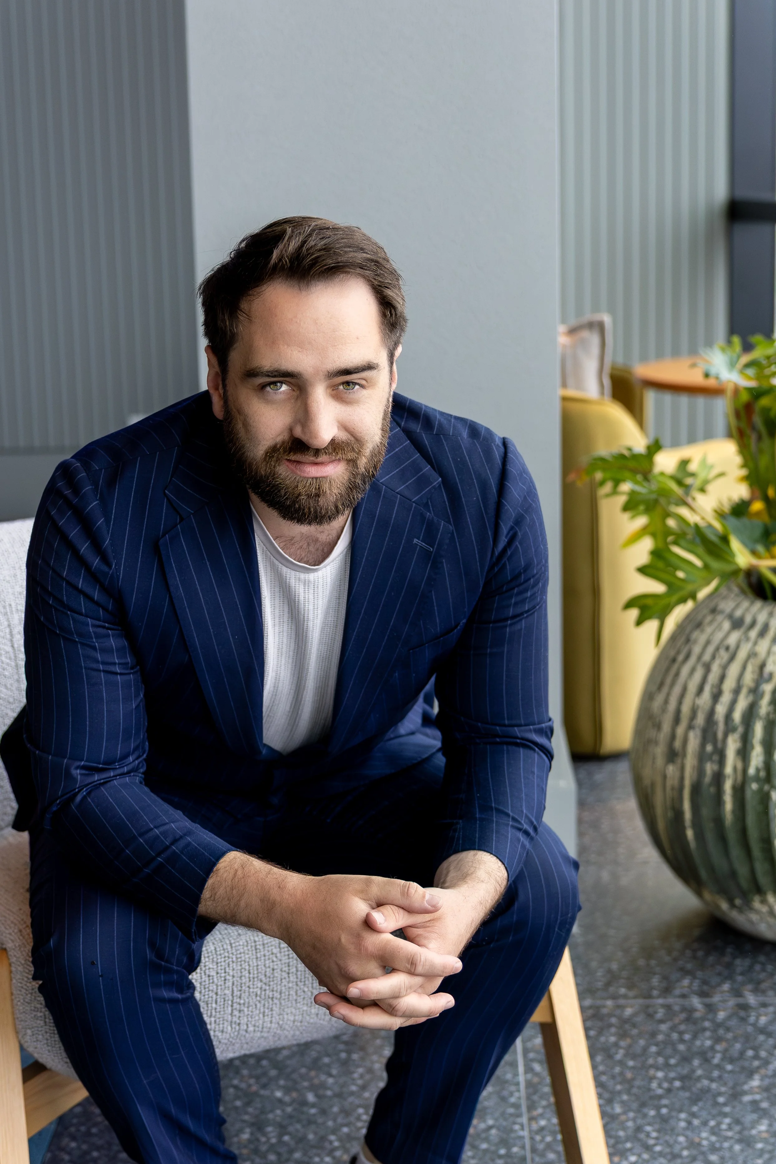 A man with a beard, wearing a dark blue pinstripe suit, sitting on a chair indoors, with plants and a small round table visible in the background.