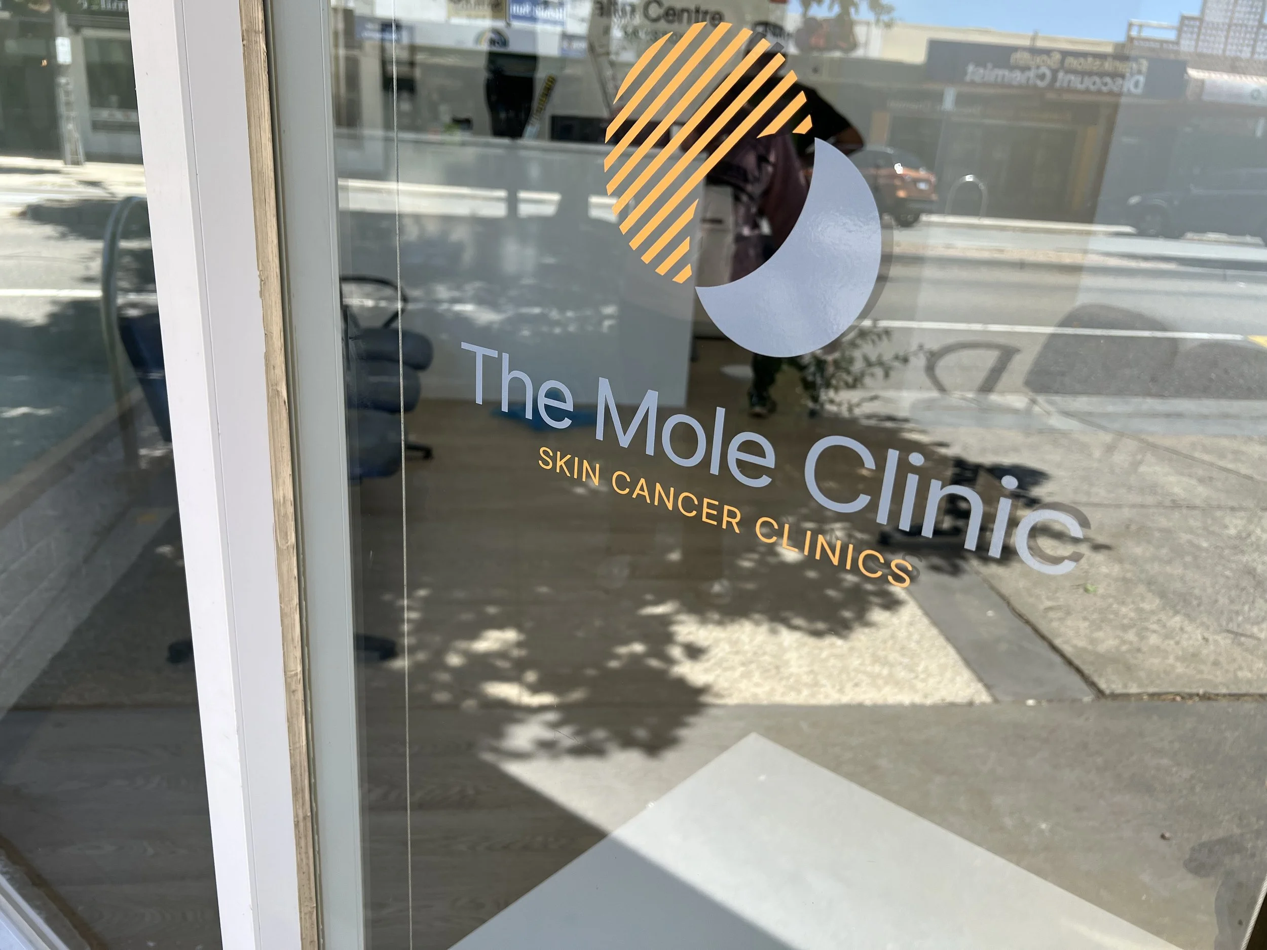 Glass door with the logo and name of The Mole Clinic, a skin cancer clinic, reflected on the glass.